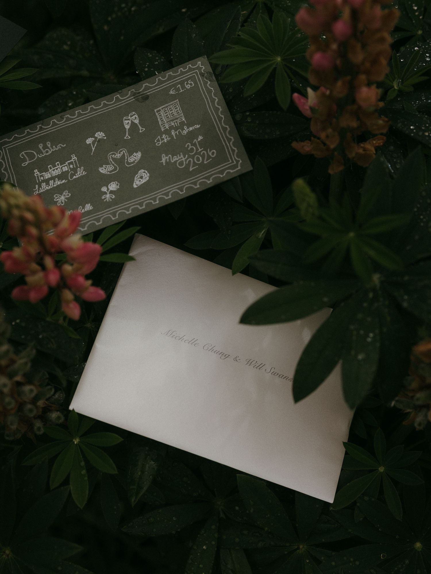 Wedding invitation and envelope resting among lush green foliage and pink wildflowers in a garden setting.