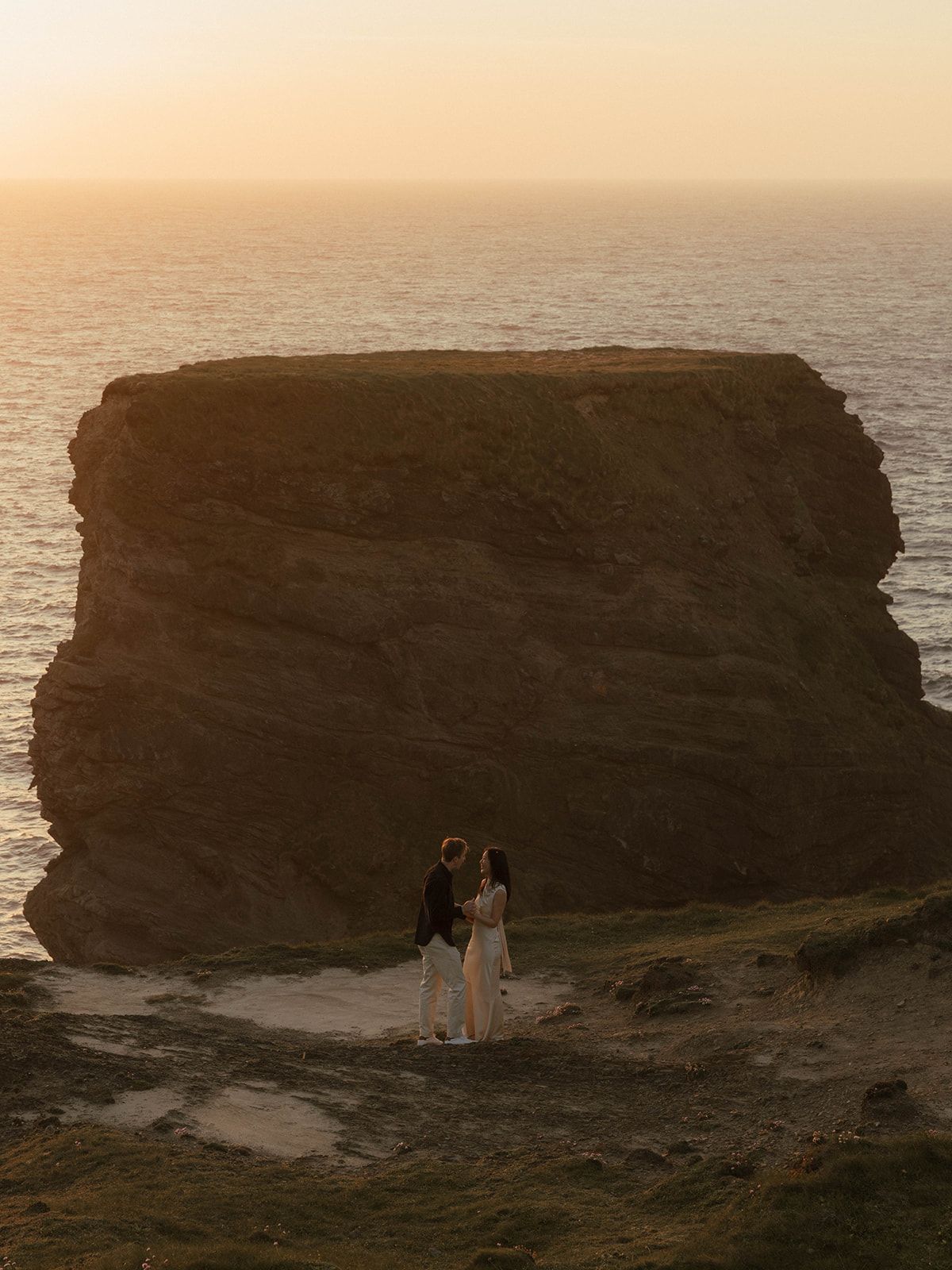 Couple standing before a massive coastal rock formation at golden sunset, overlooking the ocean.