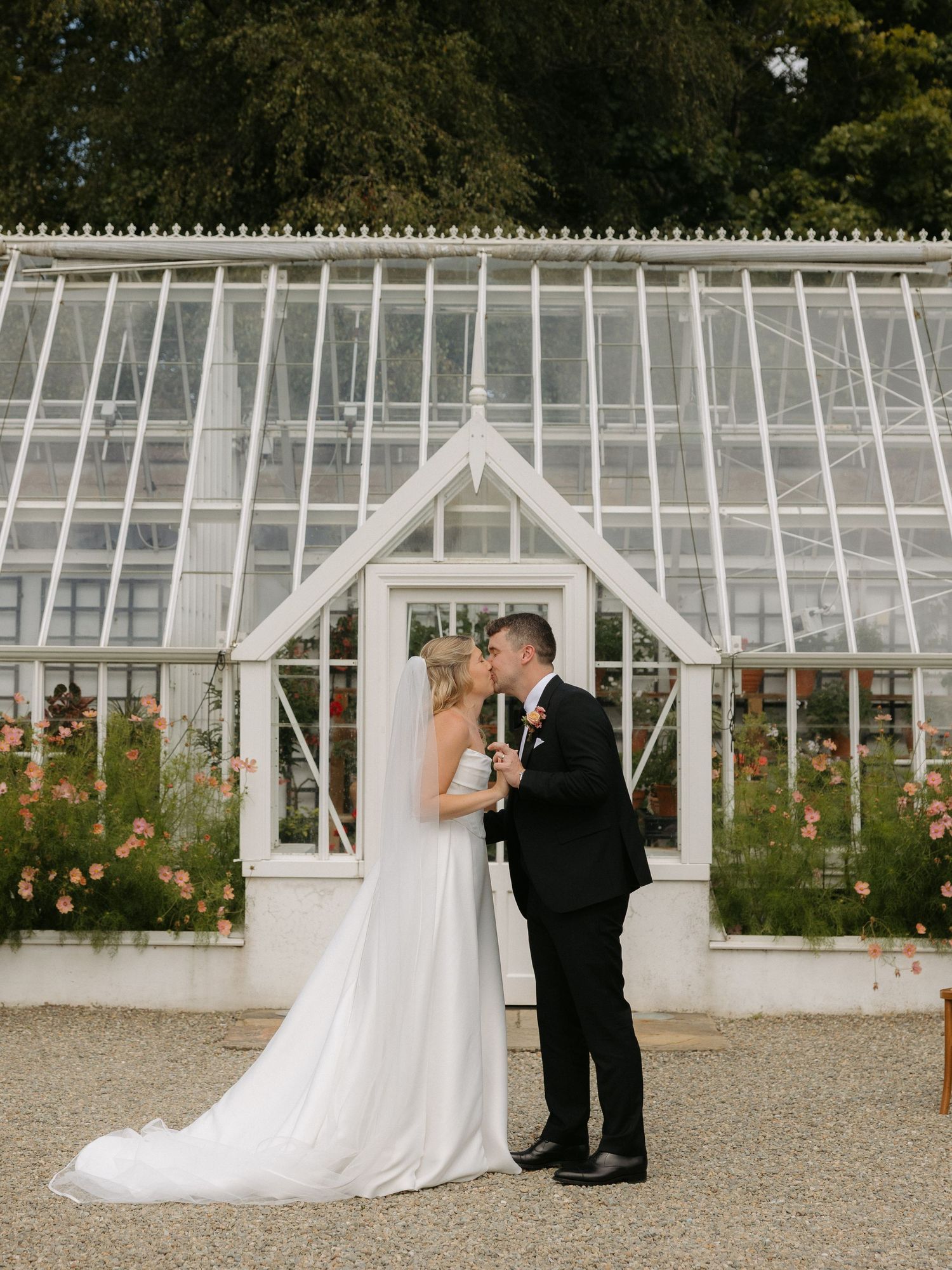A romantic moment between a couple in formal attire at a greenhouse venue with white architecture and blooming flowers.