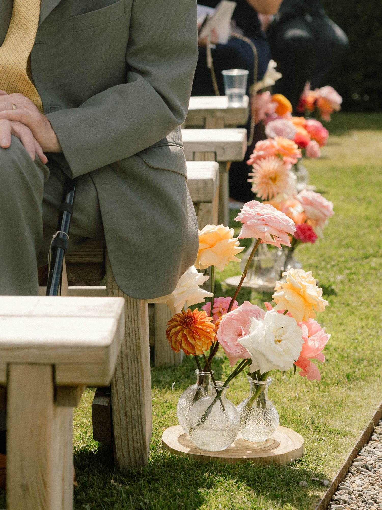 Colorful coral and peach dahlias decorate wooden benches at an outdoor garden wedding ceremony.