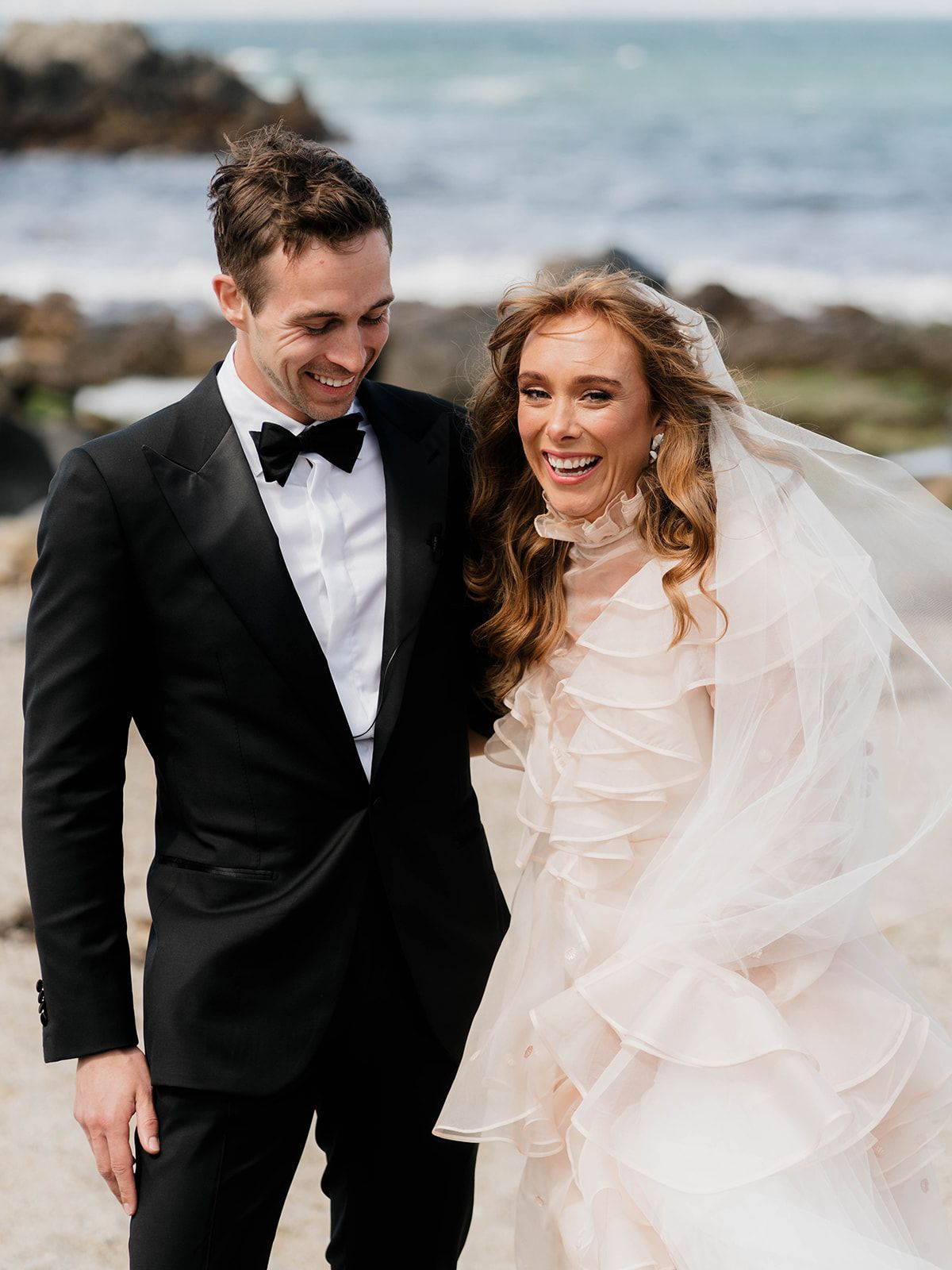 Joyful wedding couple shares a laugh on rocky beach with ocean waves behind them.