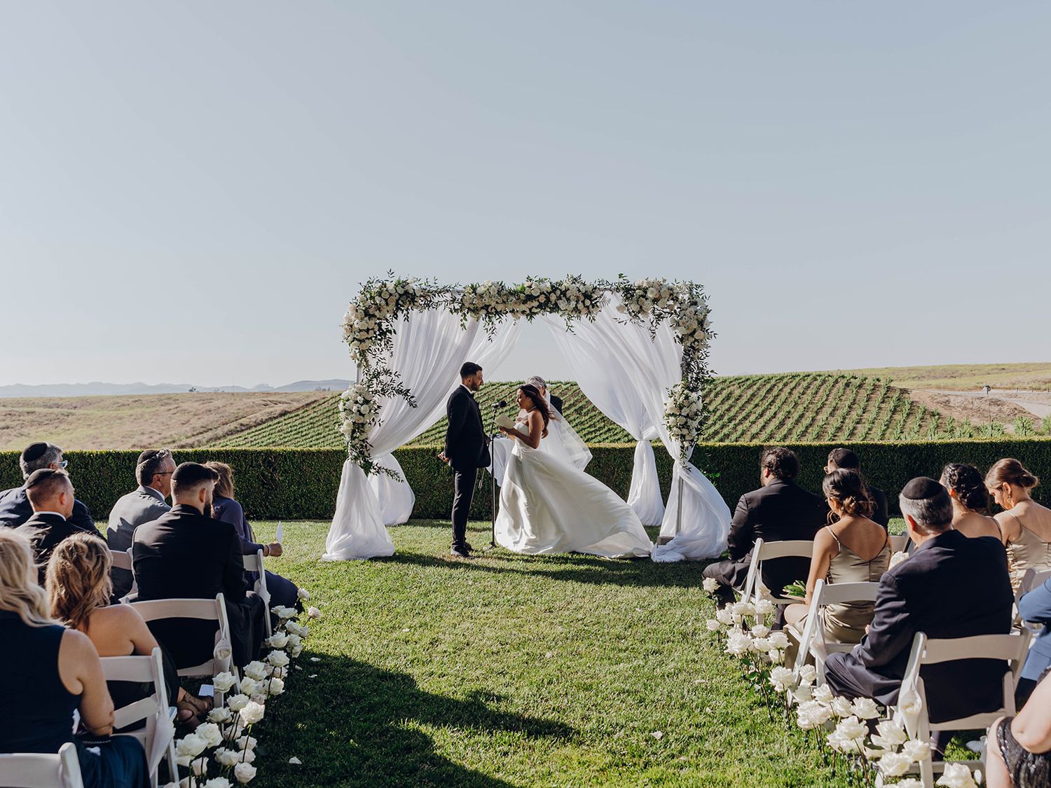 An outdoor wedding ceremony overlooking vineyard hills with floral arch and guests seated in white chairs on a grassy lawn area.