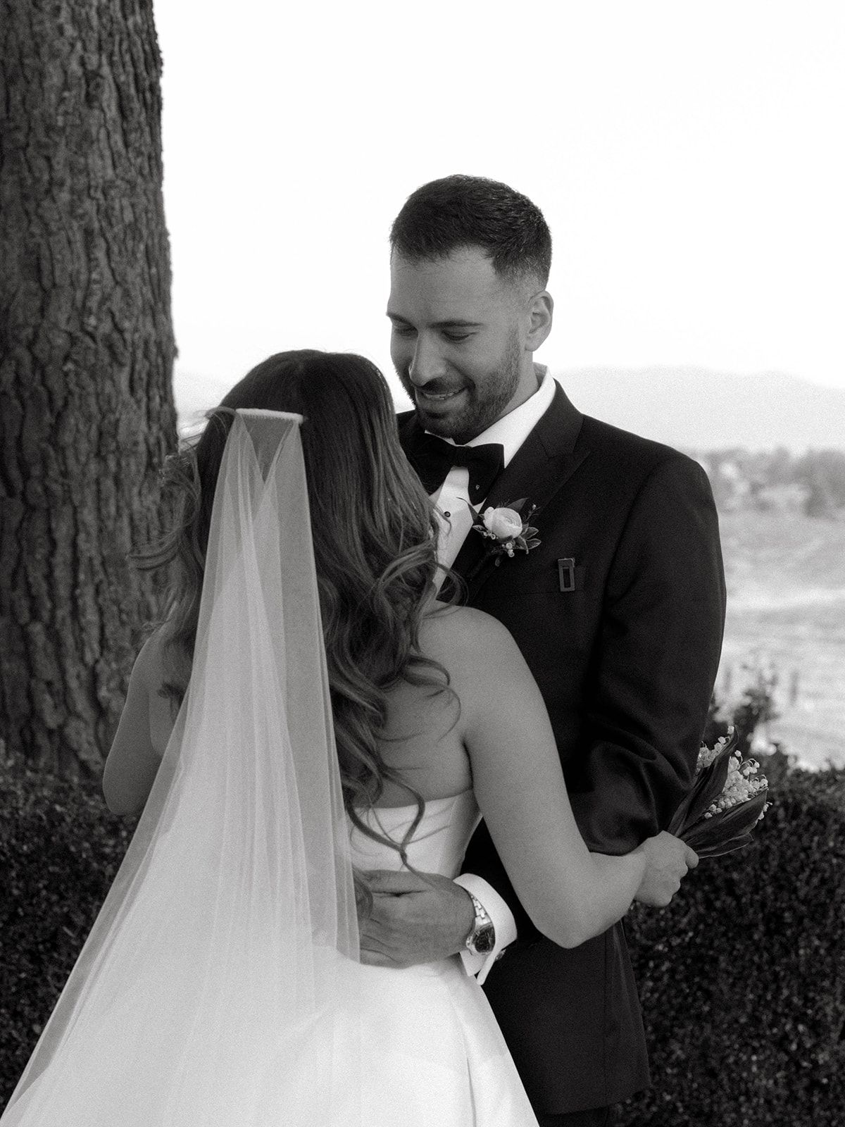 A tender black and white wedding moment captured under a tree as a couple shares an intimate embrace.