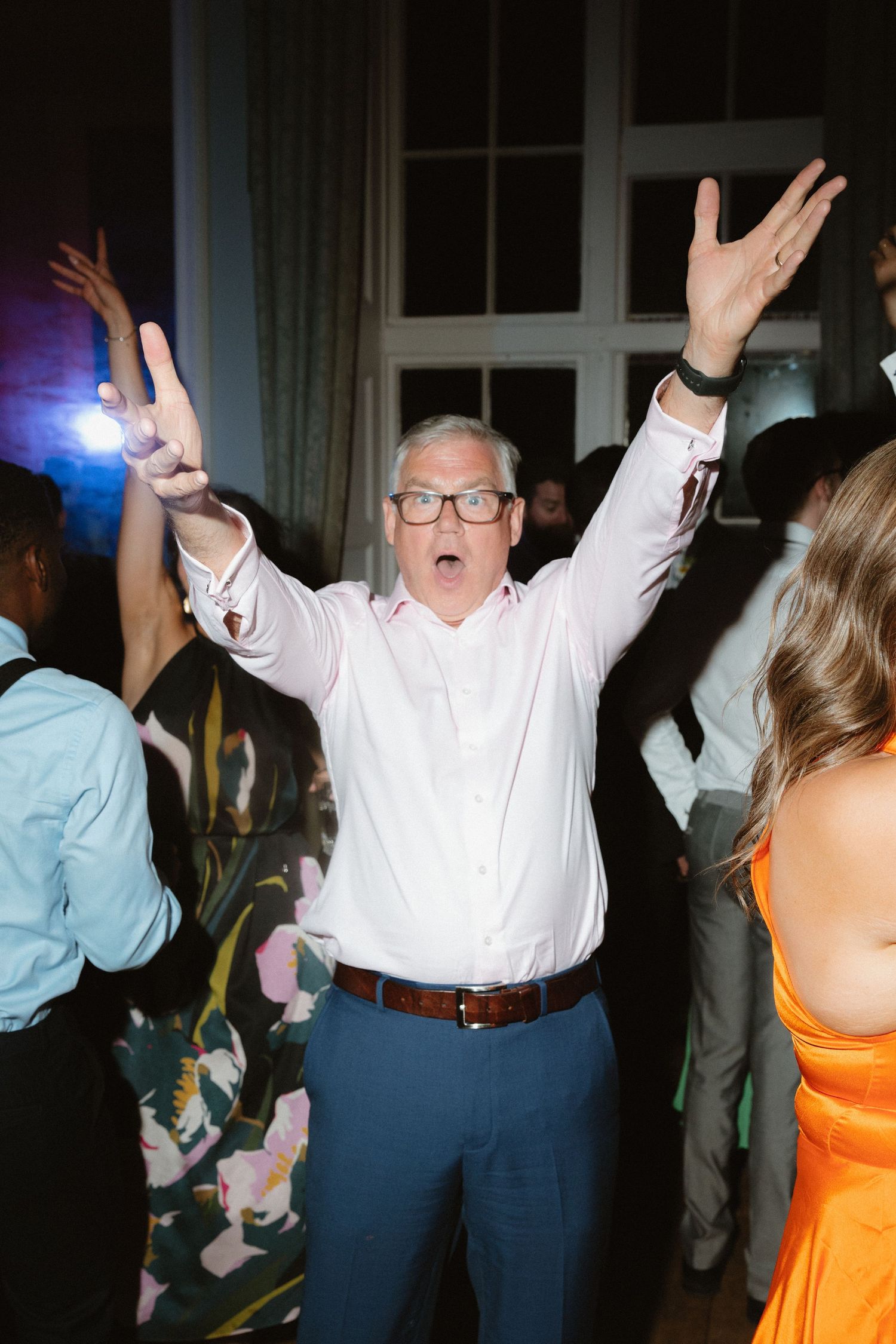 An enthusiastic guest in white shirt and glasses dances with raised arms at a wedding reception.