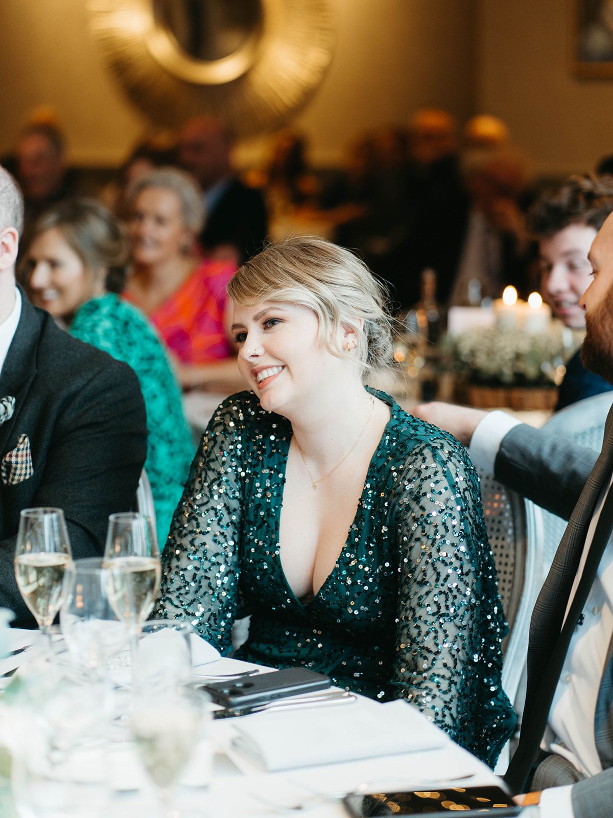 A guest in a sparkly emerald green dress sits at an elegant wedding reception dinner table with wine glasses.