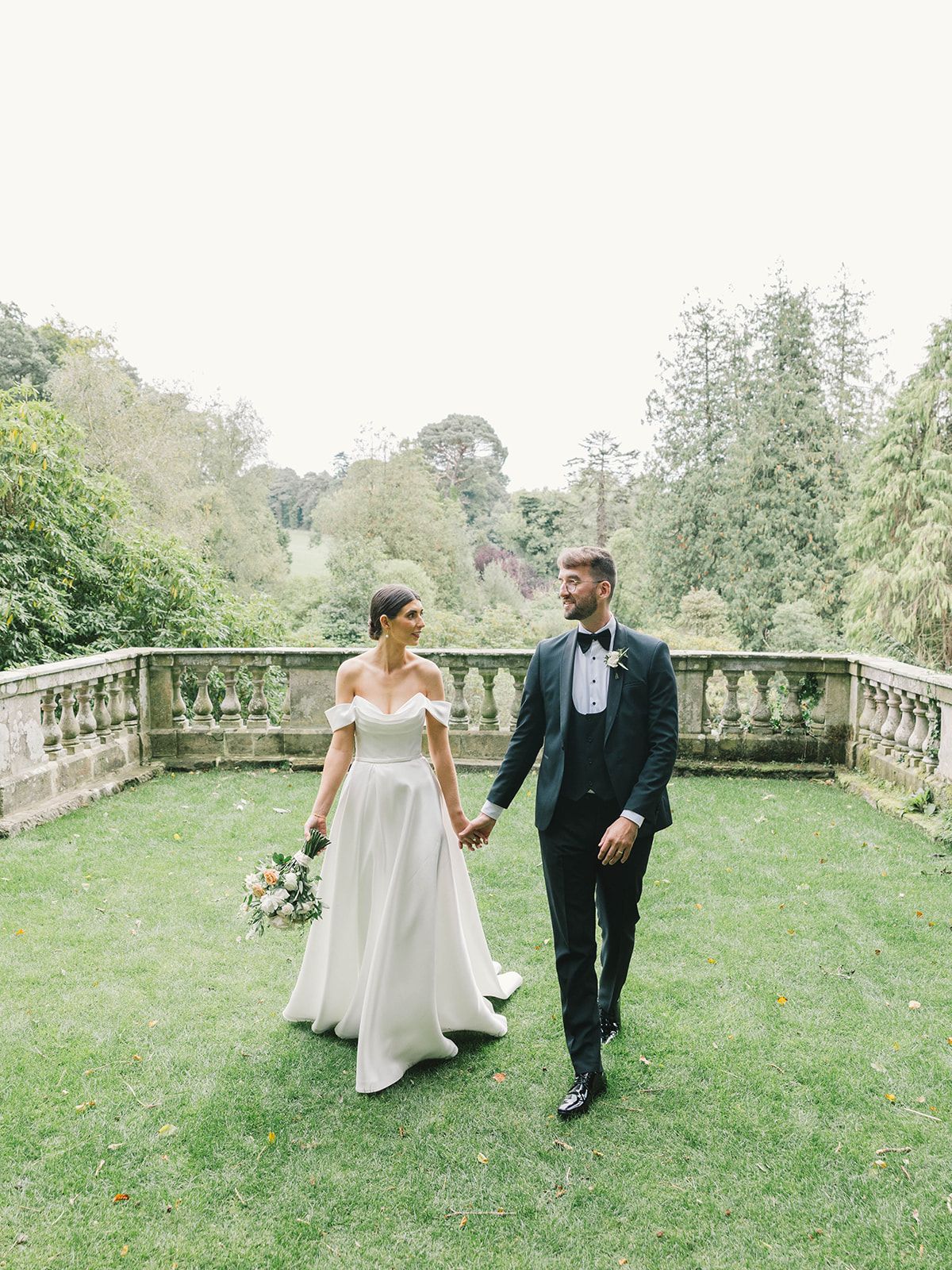 A bride and groom walk together on a beautiful garden terrace with ornate stone railings on a sunny day.