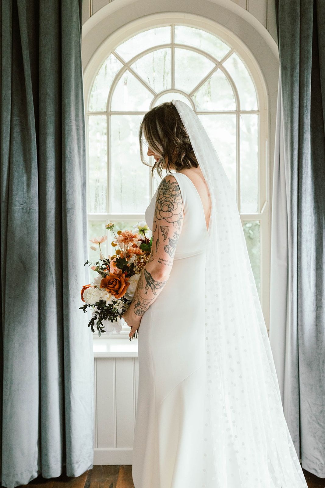 Elegant bridal portrait in front of arched window showing long sleeve lace gown, flowing veil and autumn-toned bouquet.