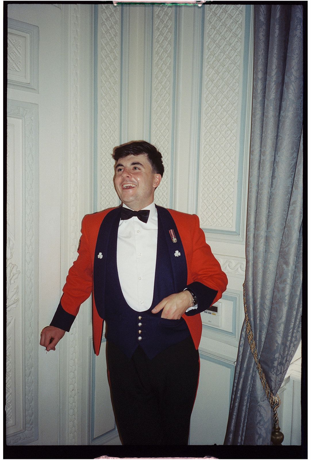 Person wearing a red tuxedo jacket and black bow tie posing against a light colored wall.
