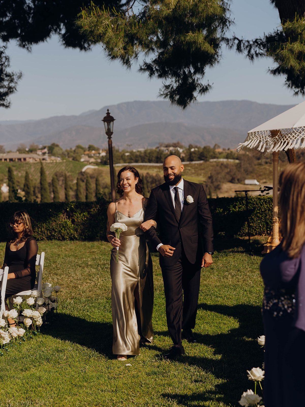 An outdoor wedding ceremony takes place on a scenic hillside estate with mountains in the background.