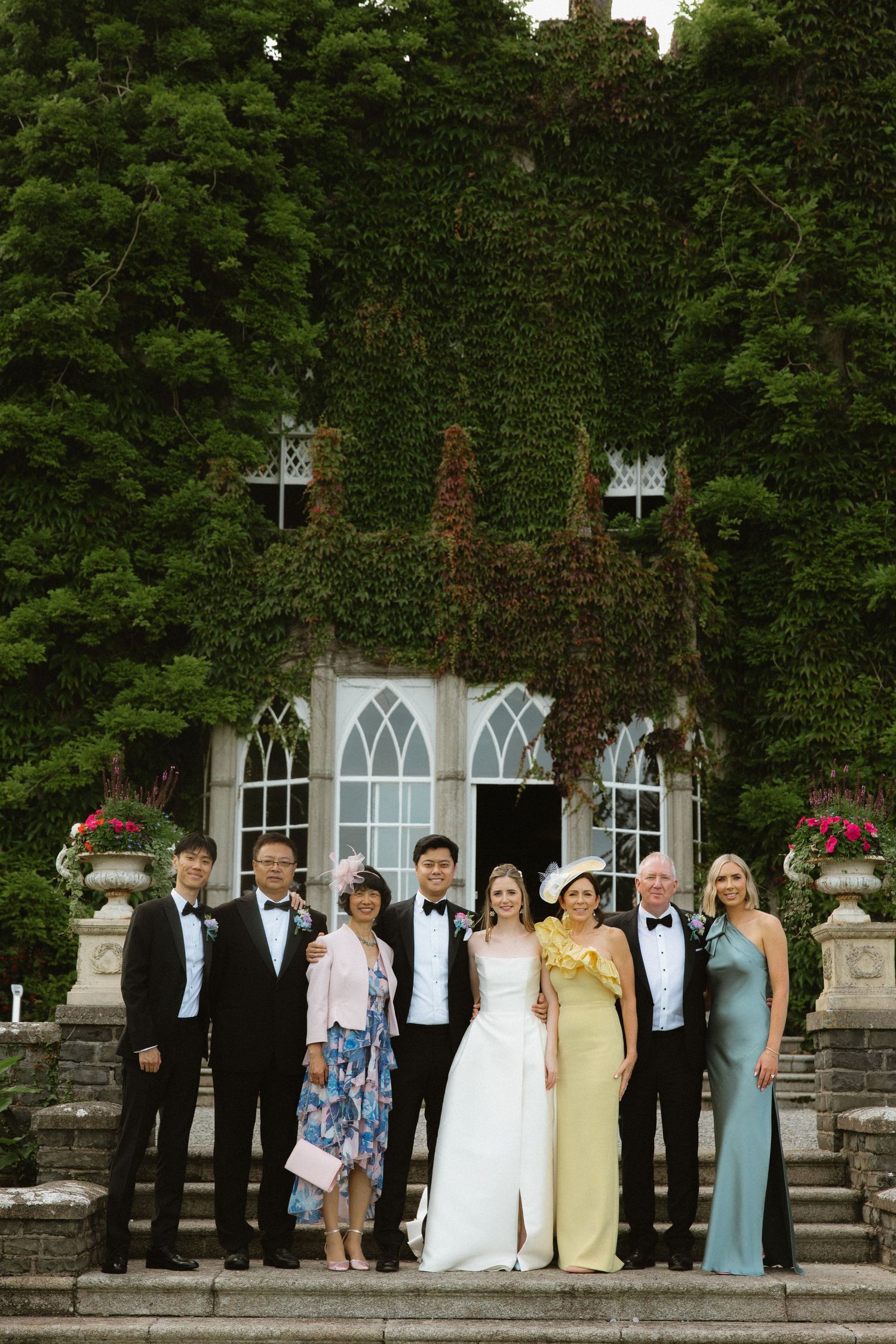 Wedding party members pose on stone steps of an ivy-covered historic building.
