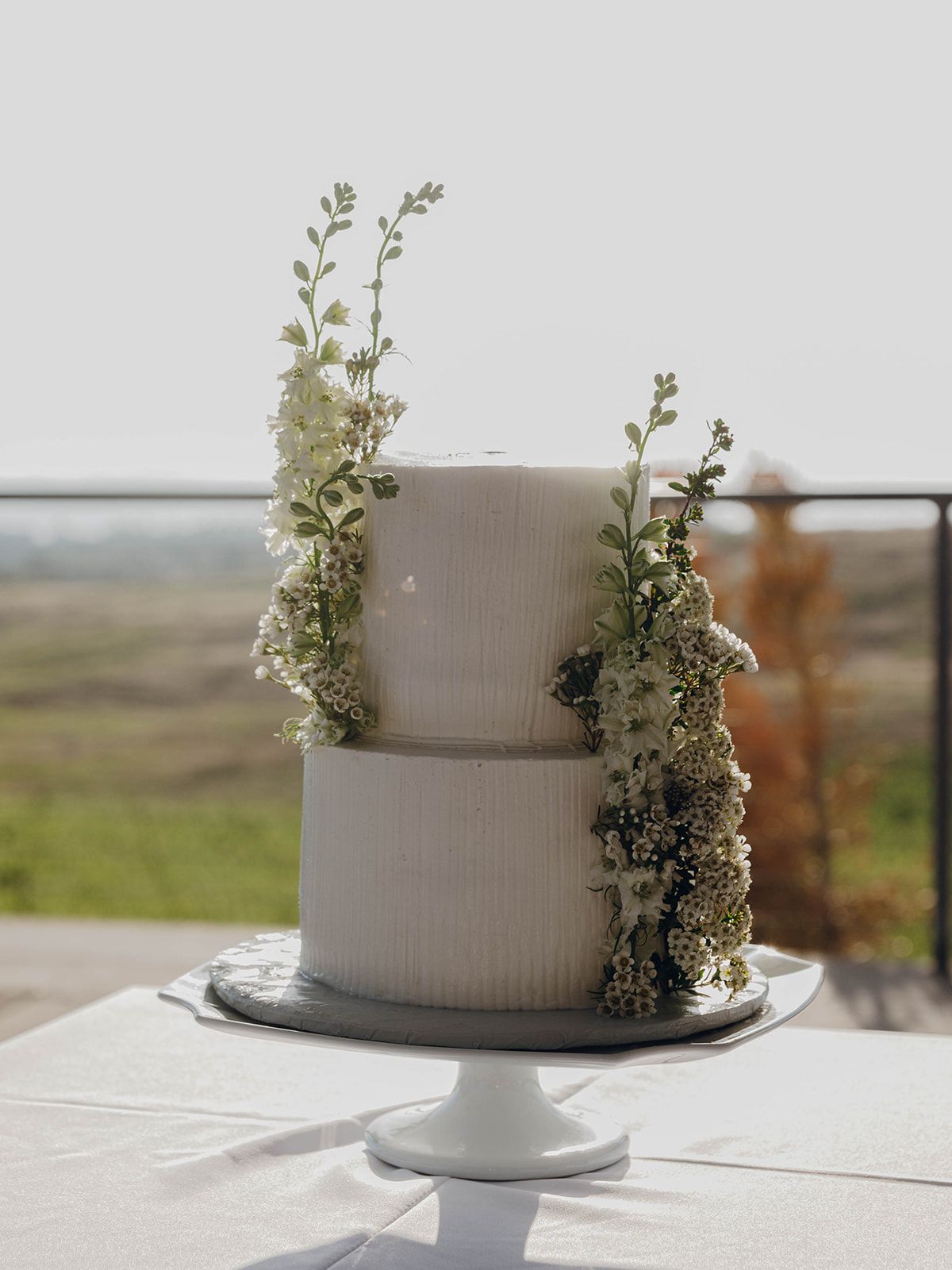 Elegant two-tier gray wedding cake decorated with delicate white flowers and greenery on a white cake stand against a scenic view.
