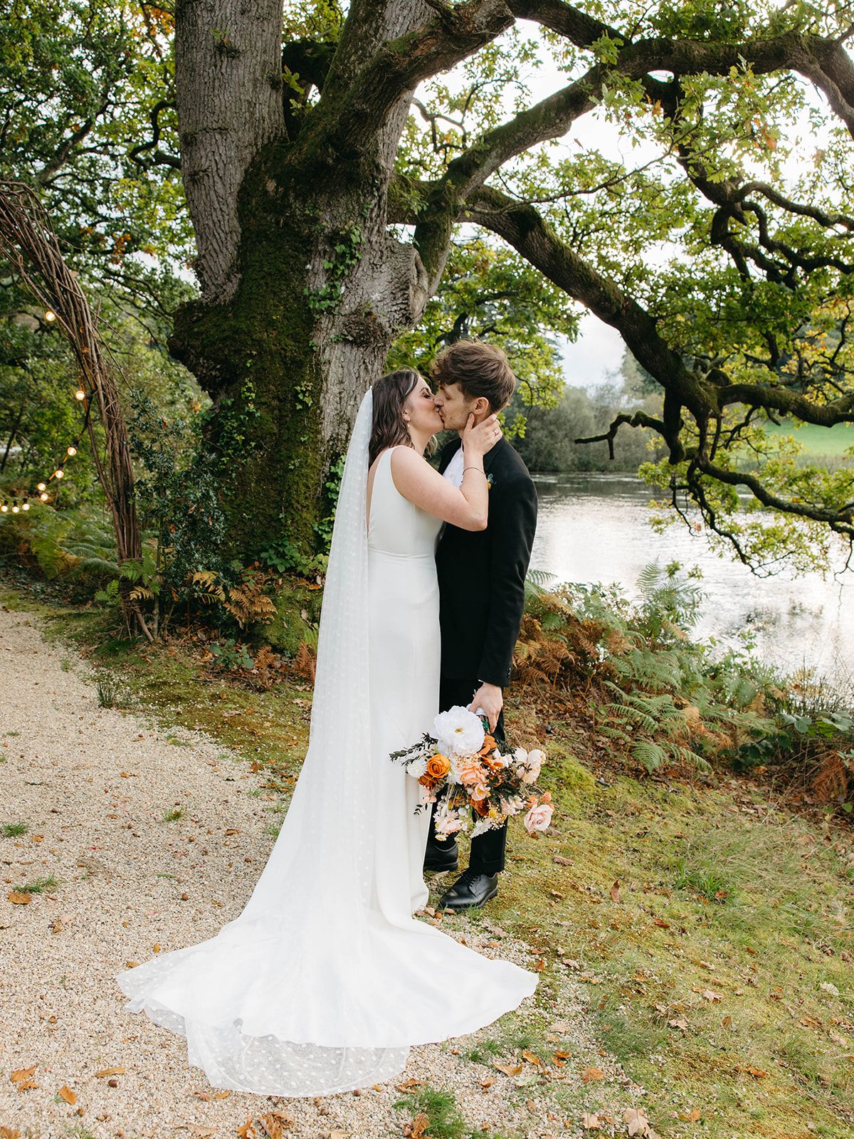 A romantic wedding couple shares a tender moment under a majestic oak tree by the water while holding a fall bouquet.