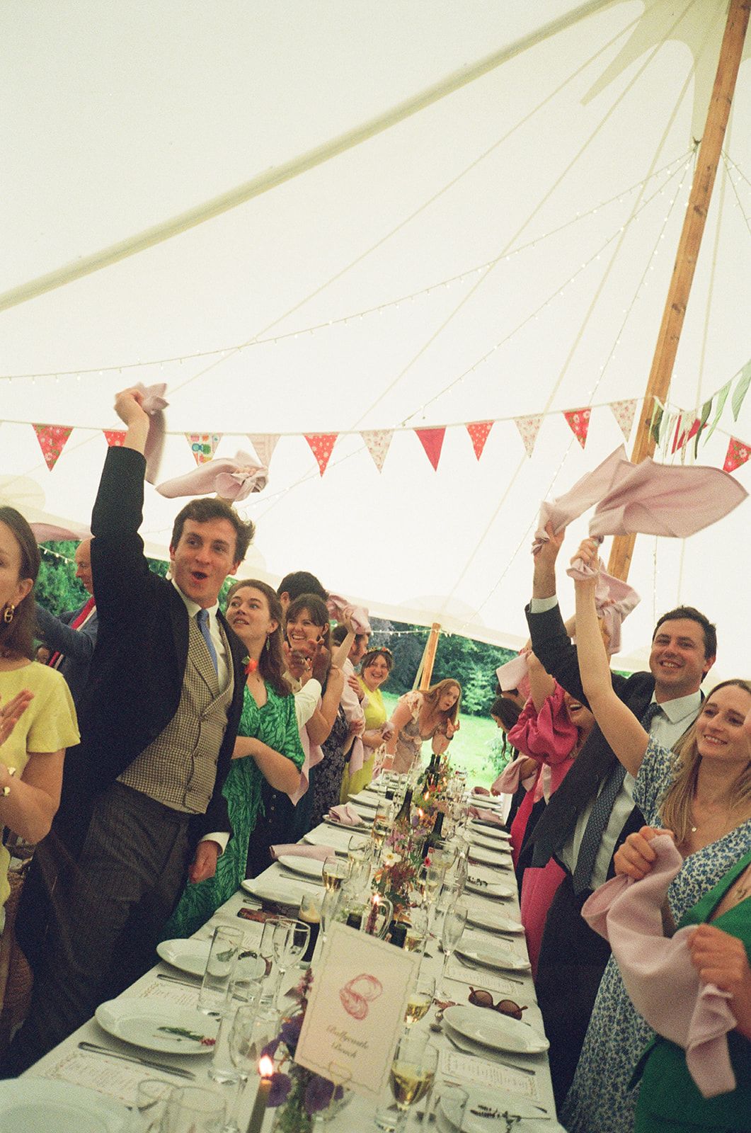 Wedding guests celebrate with raised glasses at a long banquet table under a white tent with festive bunting decorations.
