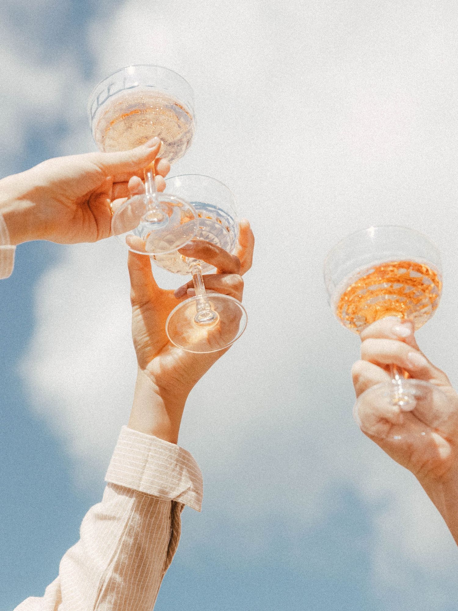 Three hands raise champagne glasses against a bright sky in a celebratory toast.