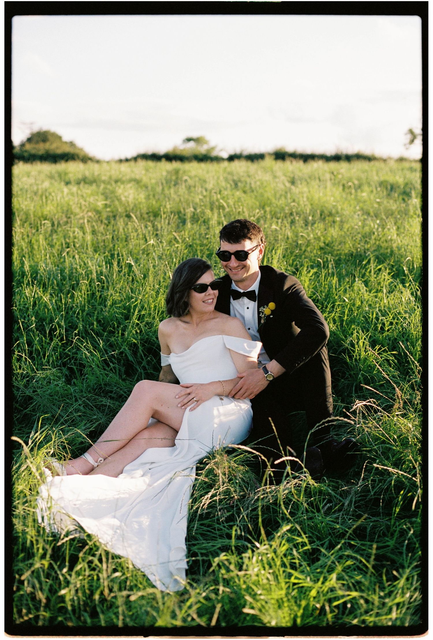 Bride and groom sitting in lush green field wearing sunglasses, sharing a romantic moment on their wedding day.