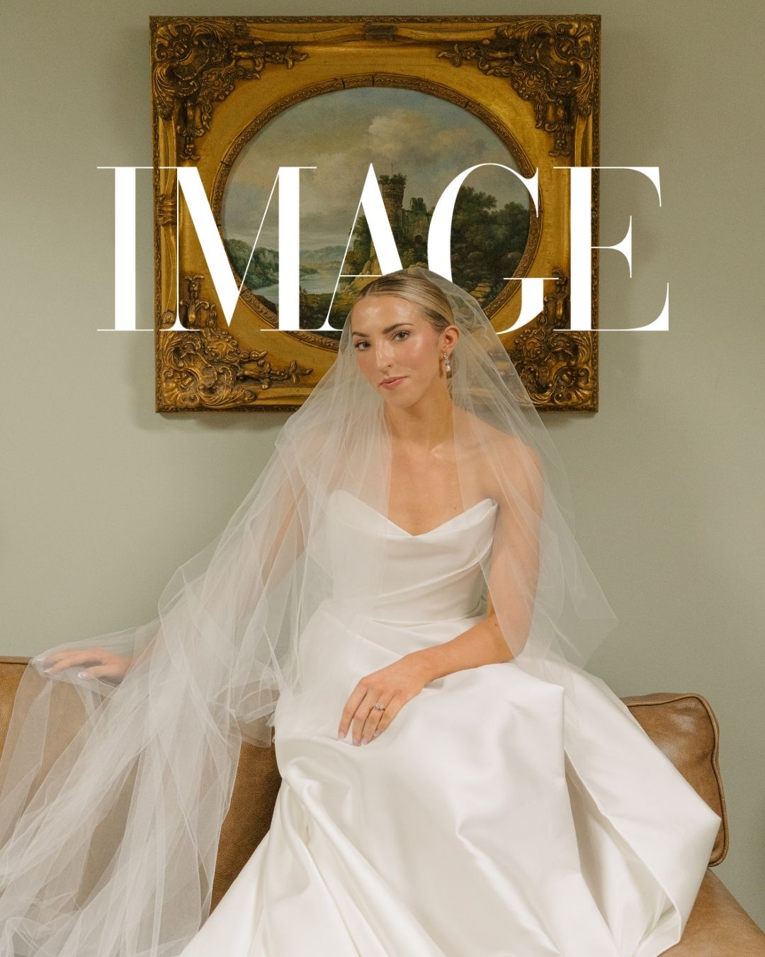 Elegant bride in a strapless white satin gown with a flowing veil, seated on a sofa before a ornate gold-framed painting.