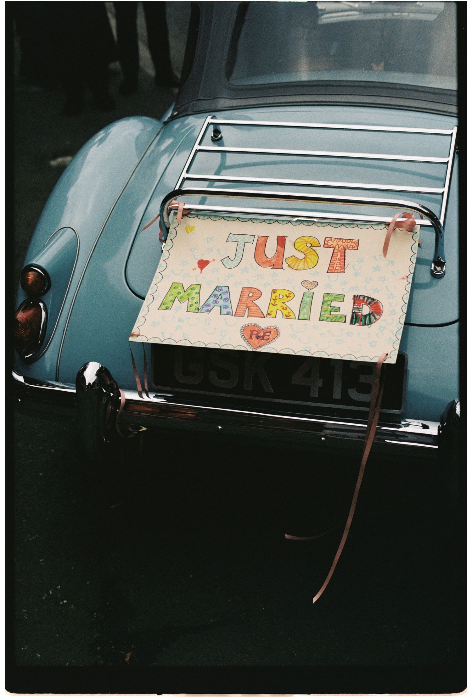 A colorful Just Married sign displayed on the back of a vintage blue car with license plate OSR 413.