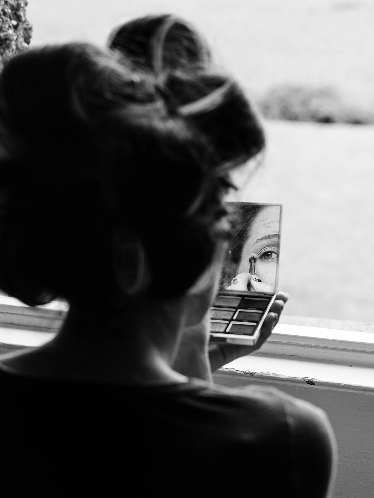 A silhouetted figure applies makeup while looking in a compact mirror in a black and white artistic photograph.