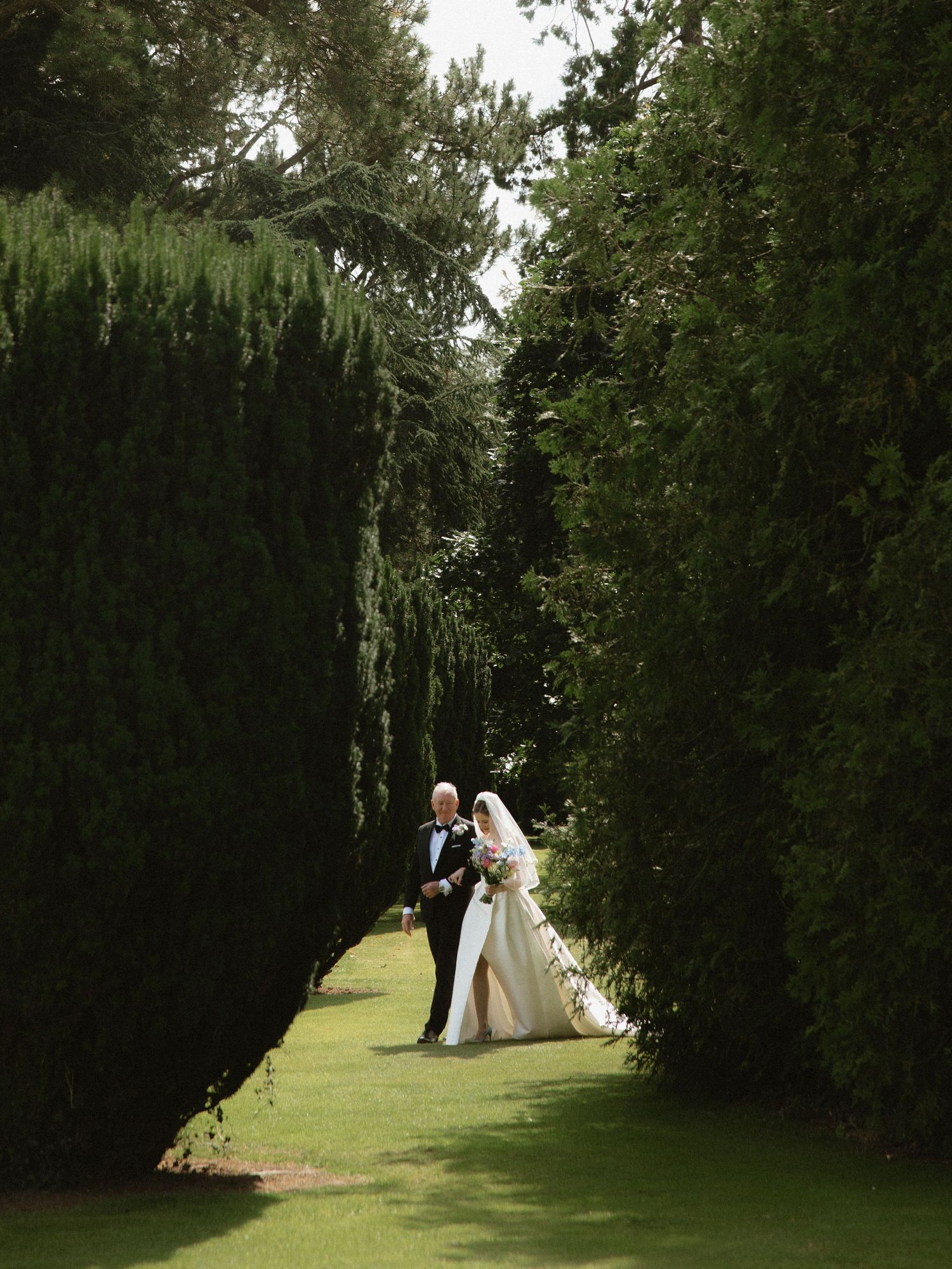 A wedding couple strolls through a manicured garden path lined with tall hedges.