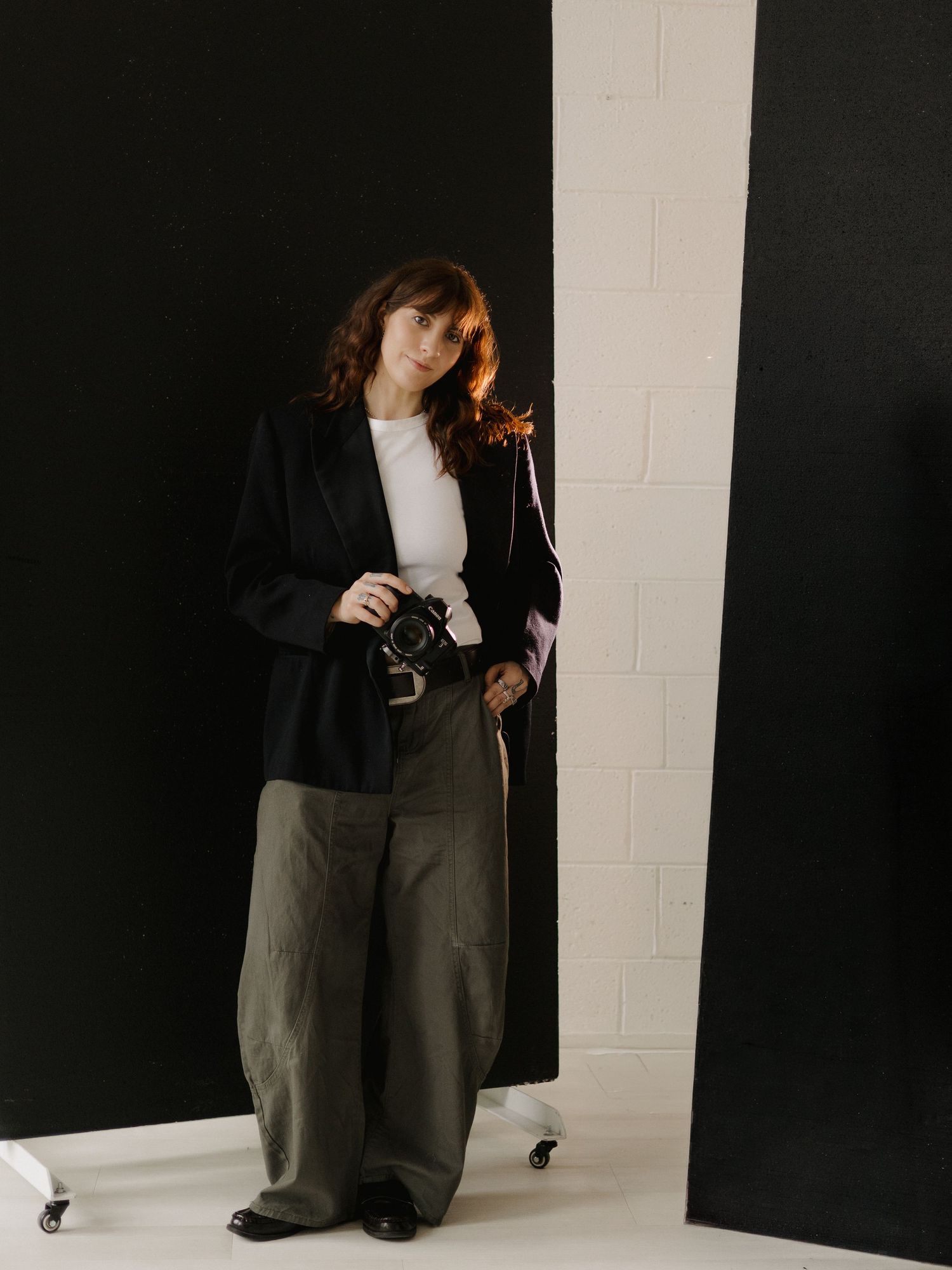 Woman in black blazer and wide-leg pants holding a camera, standing in a photography studio.