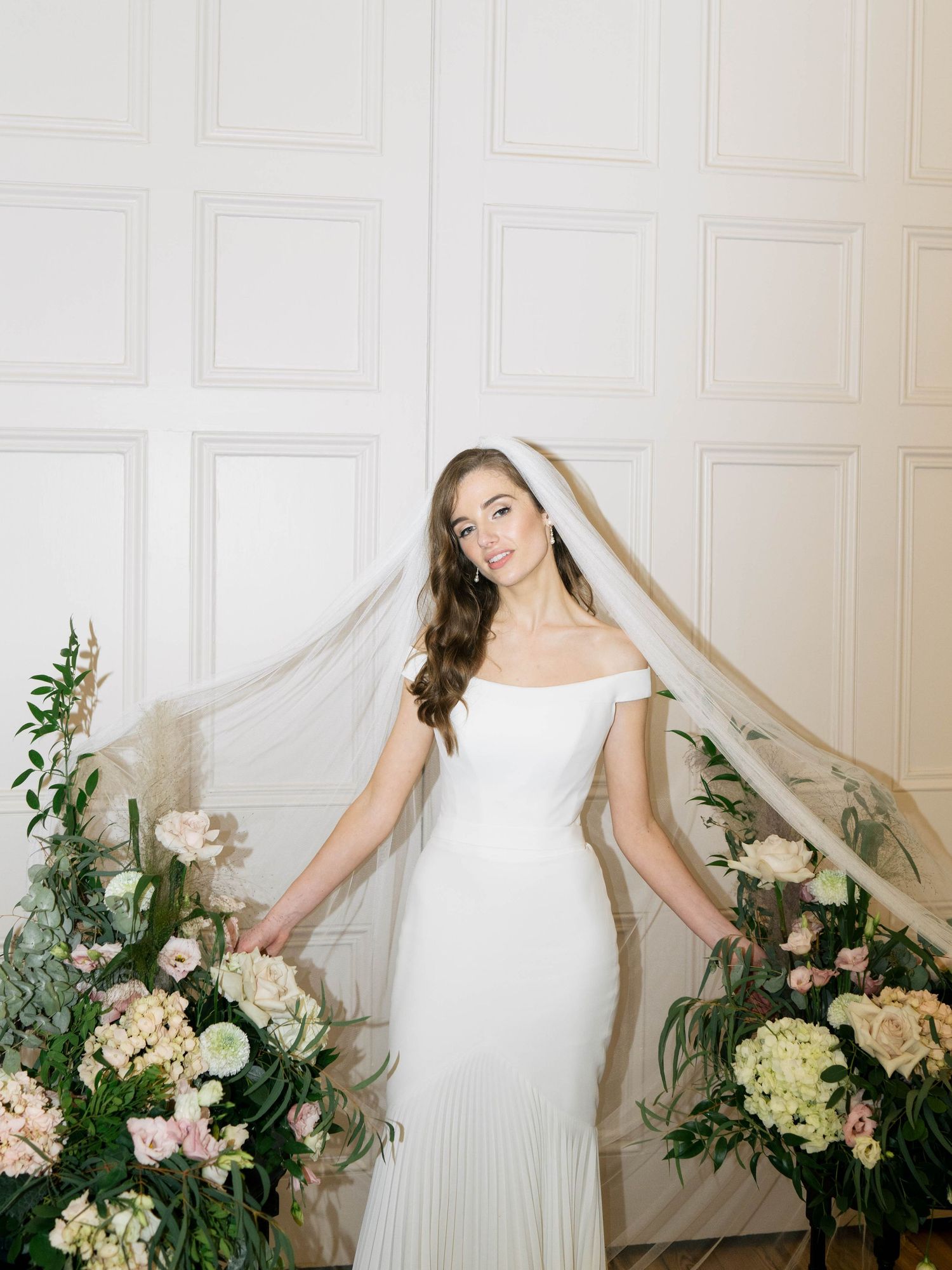 Bride in elegant white wedding gown poses gracefully against white paneled wall with romantic floral arrangements.