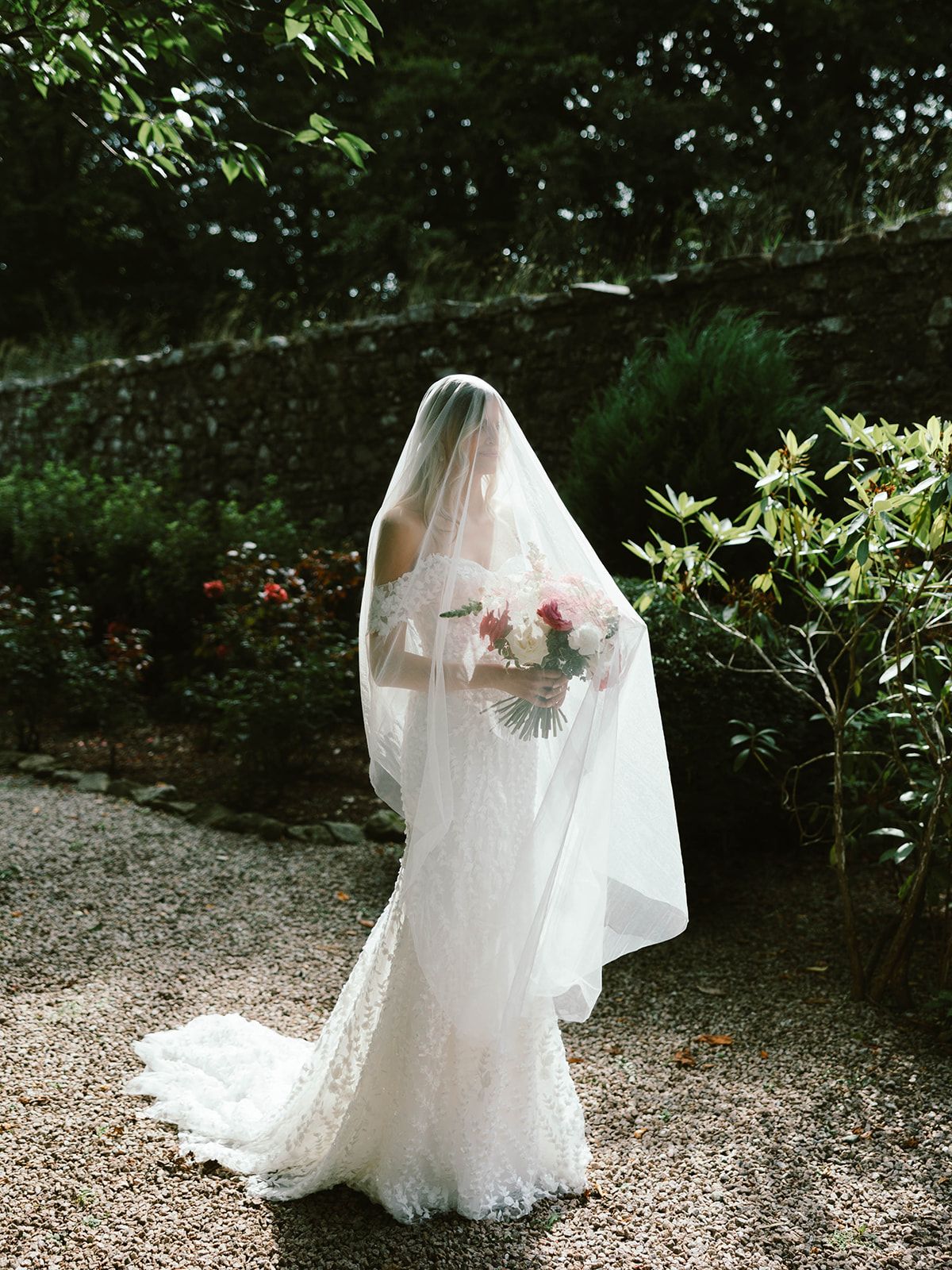 A bride in a flowing white wedding gown and veil stands in a sunlit garden path surrounded by lush greenery.