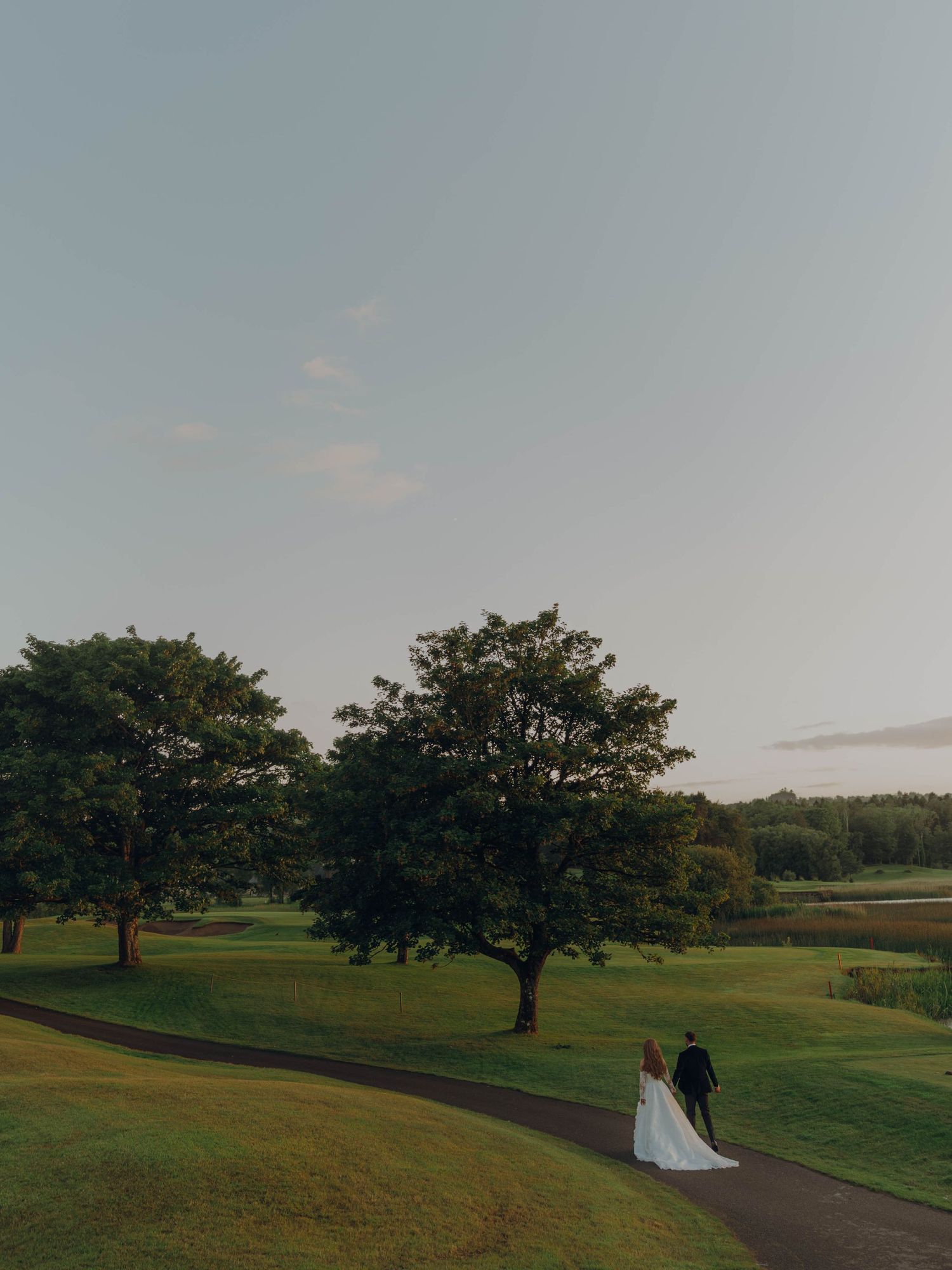 A scenic golf course landscape with rolling green hills, mature trees, and a winding cart path under a cloudy sky.