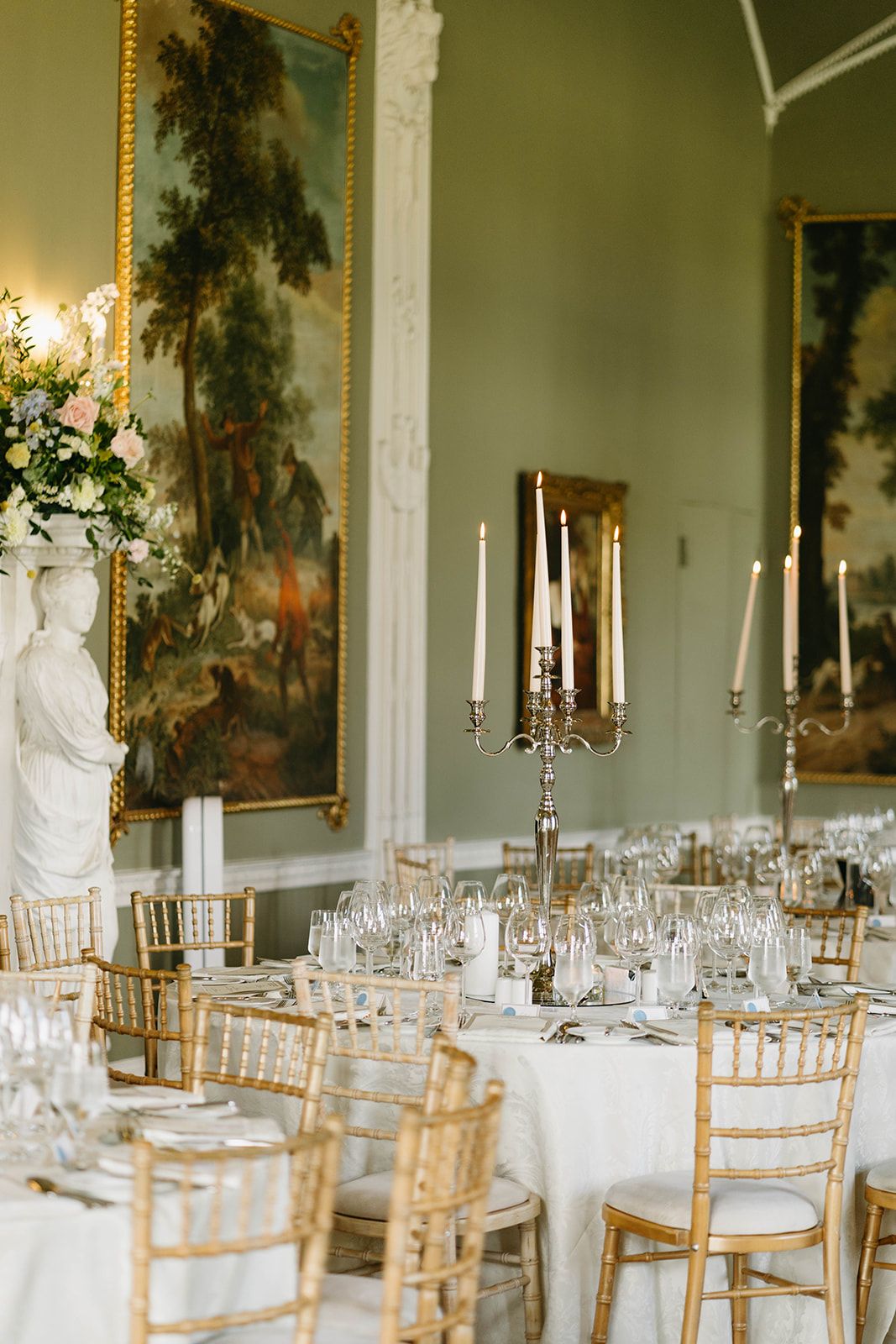 Elegant dining room setup with white tablecloths, gold chiavari chairs, and classical artwork on mint green walls.