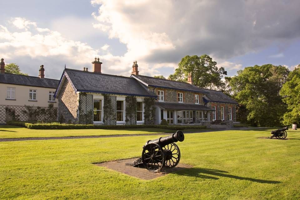 Historic stone cottages with traditional architecture line a grassy field where an antique cannon sits on display.