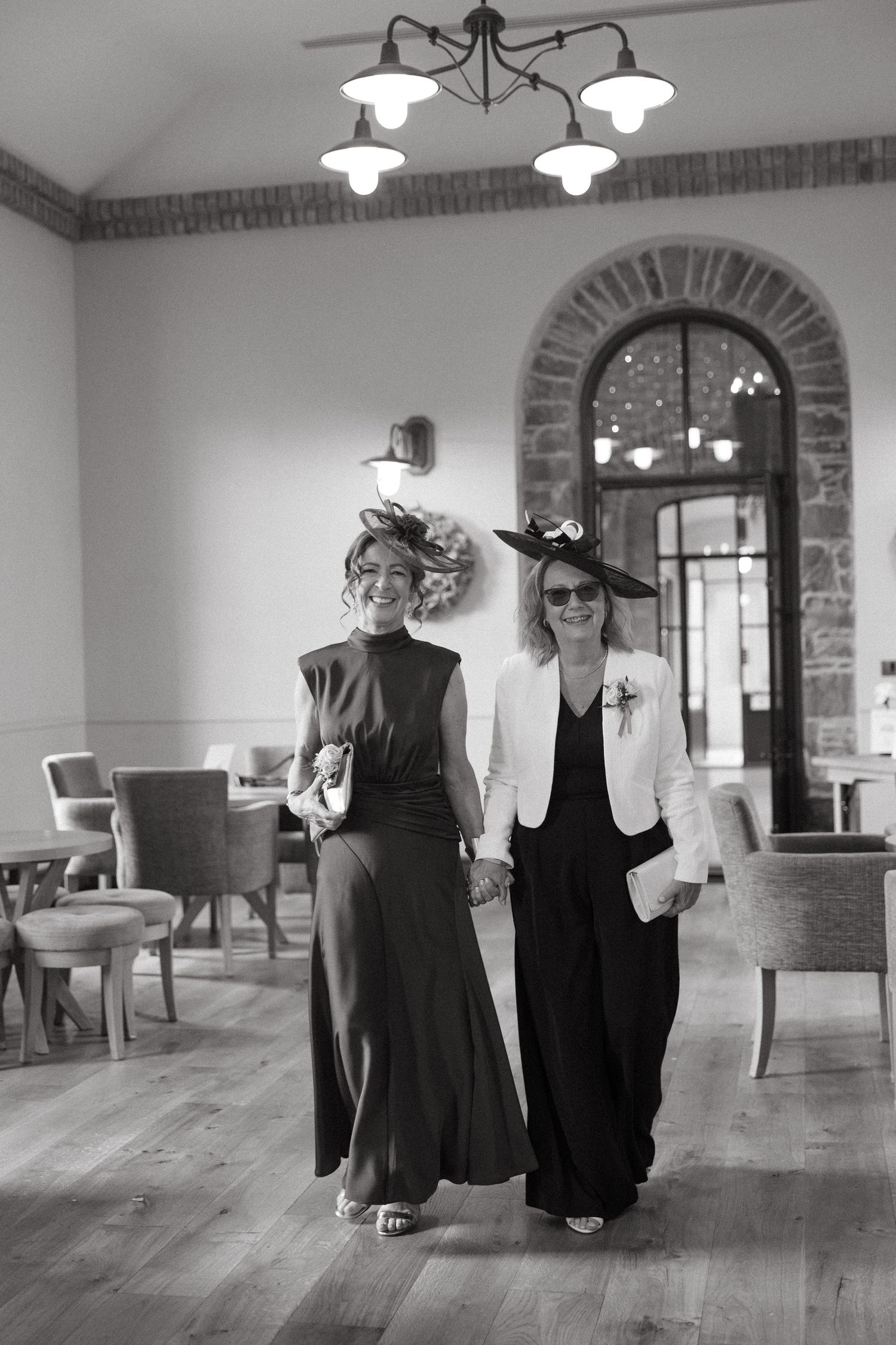 Two figures in dark clothing pose together in a bright interior space with wooden floors and arched brick details.