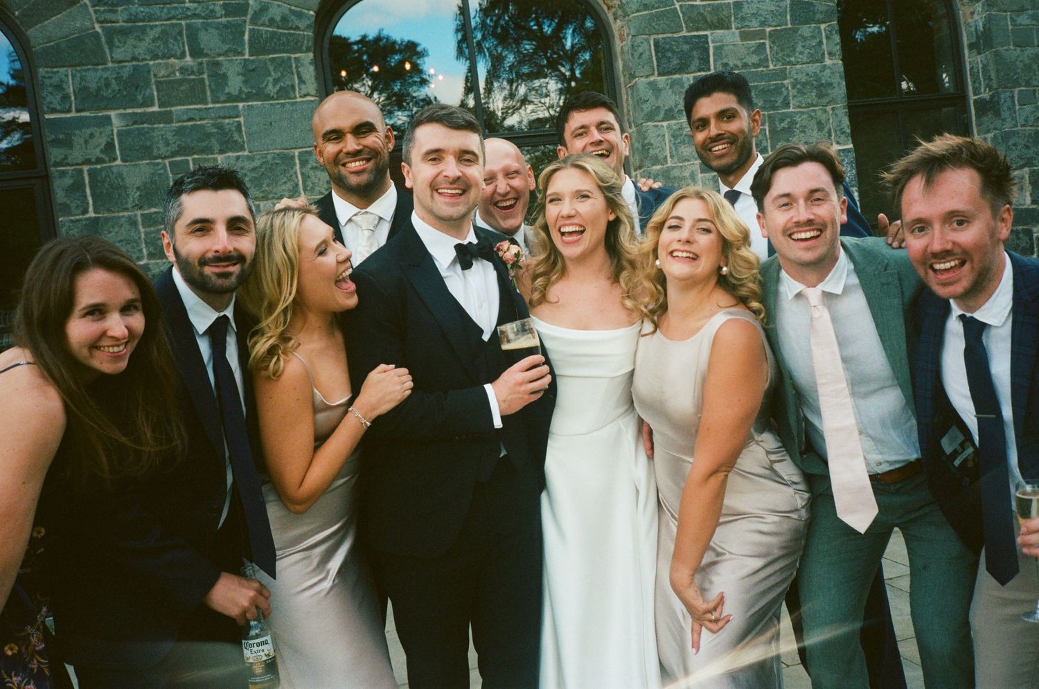 Wedding party members pose together outside a stone building during a celebration.