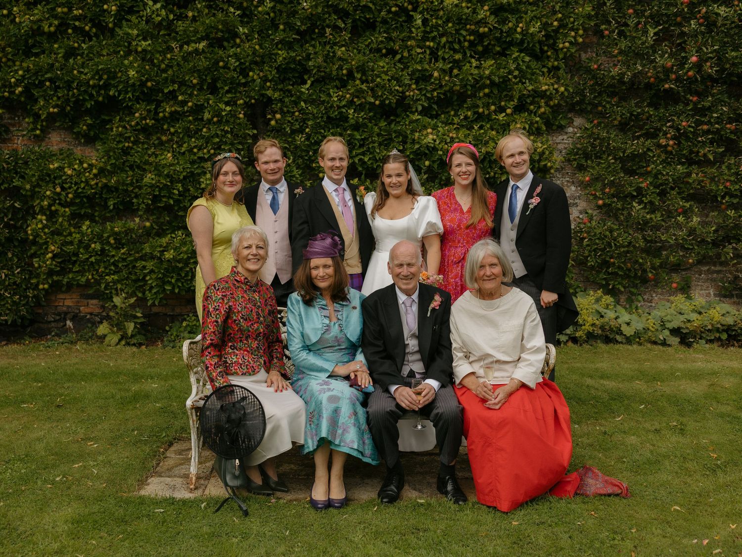 A formal group photo taken outdoors against a lush green hedge backdrop in a garden setting.
