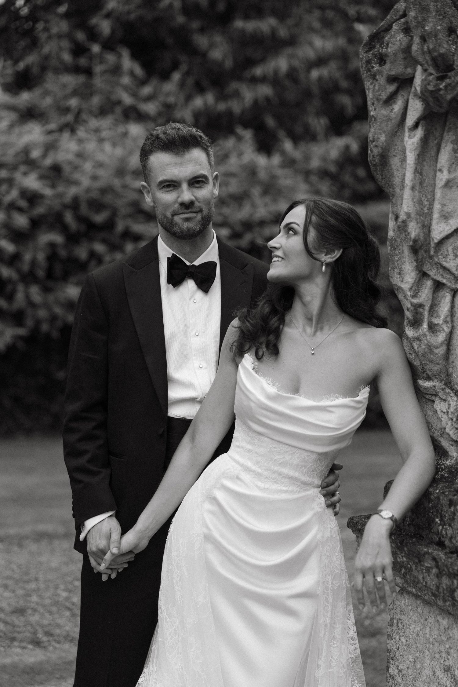 Black and white photo of a bride in strapless gown and groom in tuxedo holding hands outdoors.