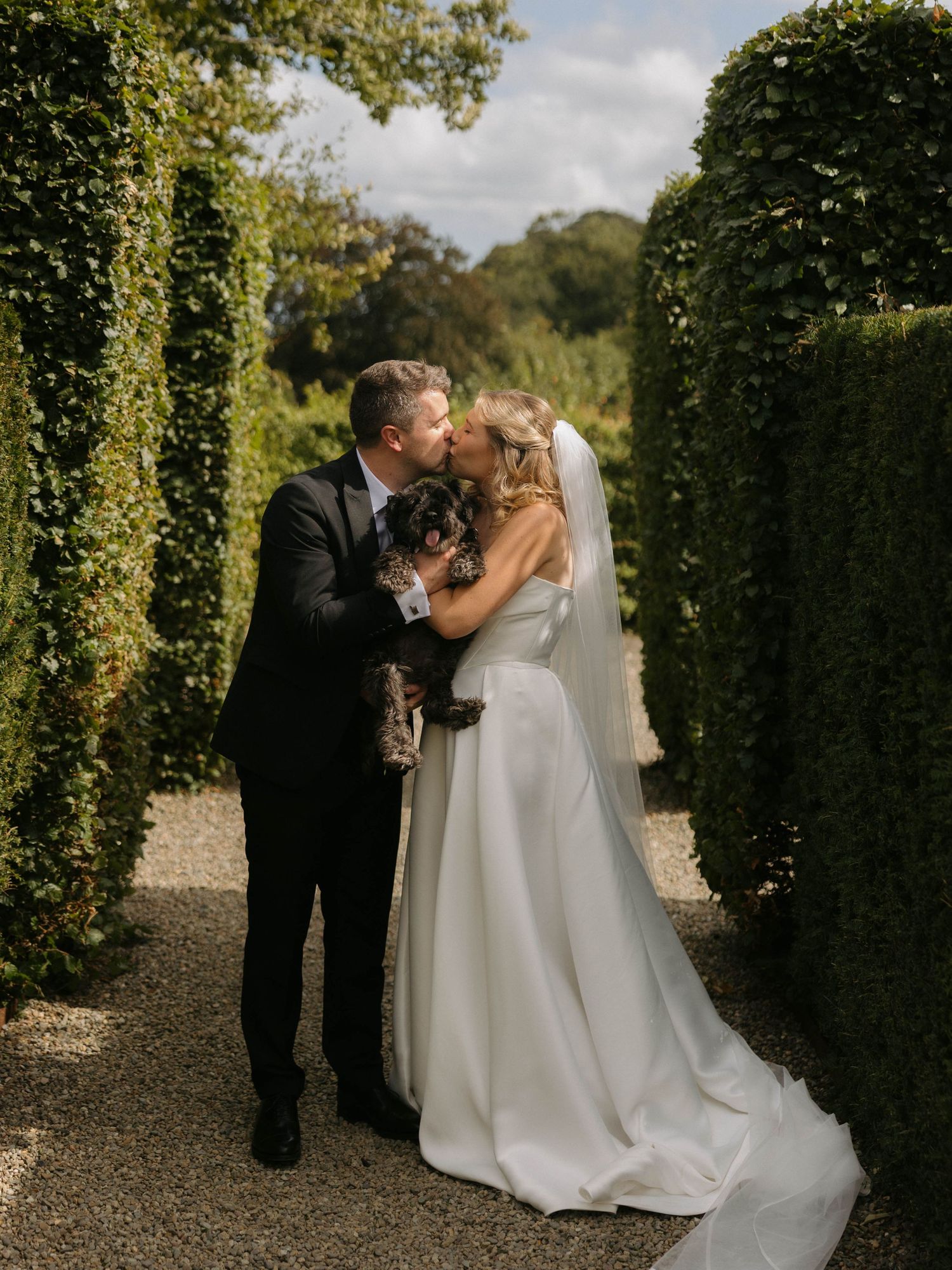 A romantic wedding couple shares intimate moments in a garden pathway lined with tall green hedges.