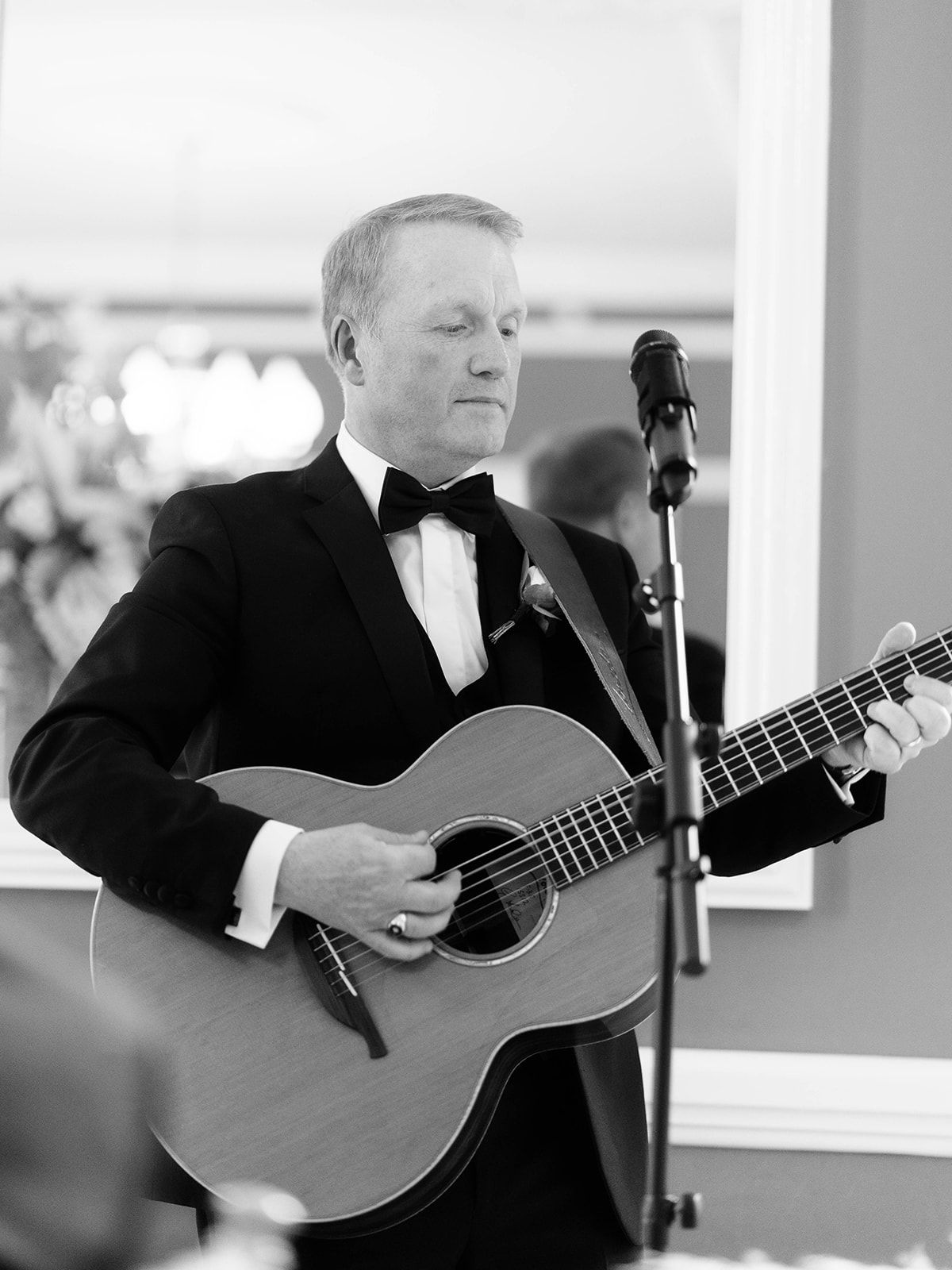 Performer in black tuxedo and bow tie plays acoustic guitar next to microphone stand in black and white photograph.