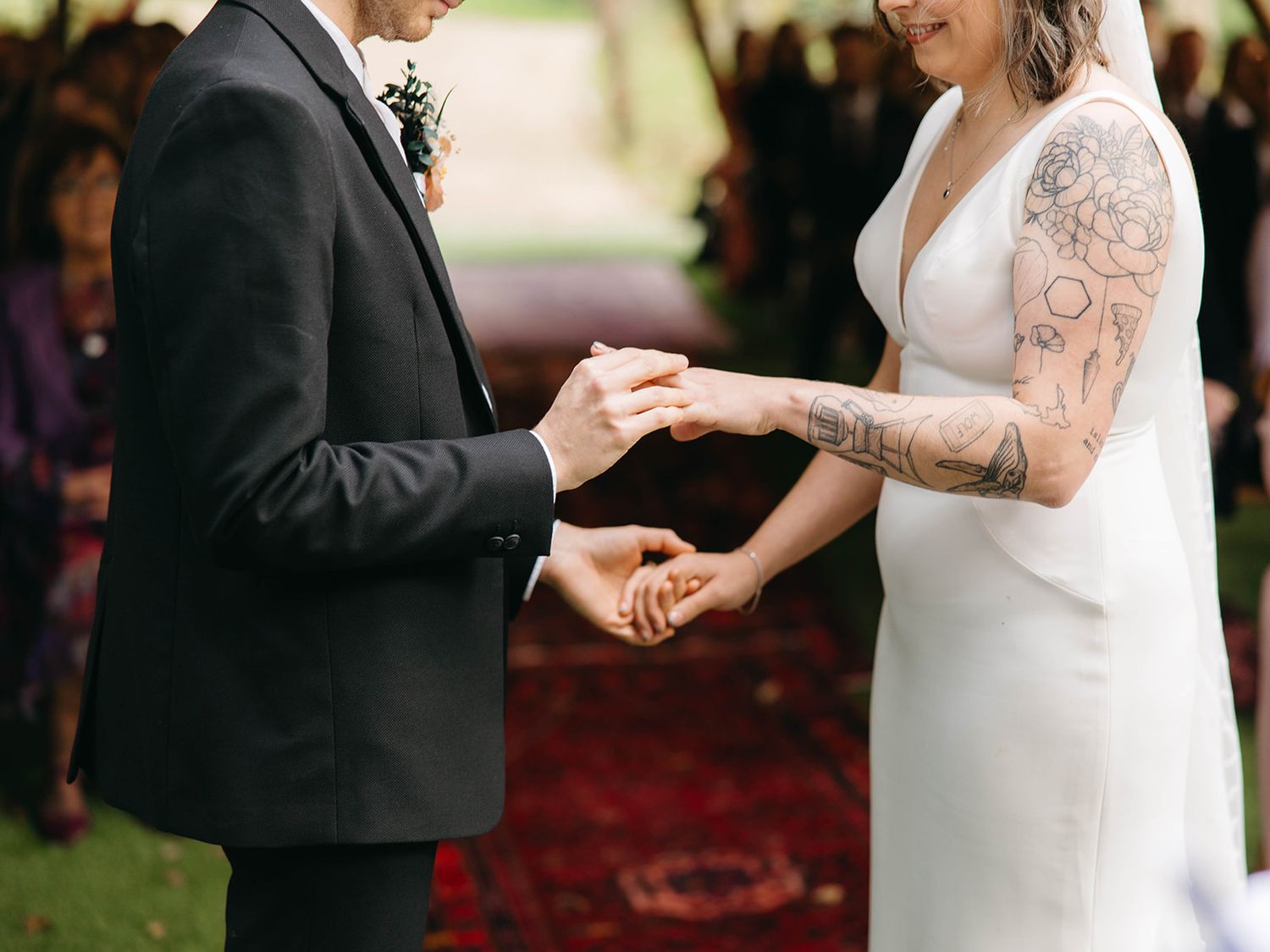 A couple exchanges wedding rings during an outdoor ceremony, with the bride wearing a white dress with lace sleeves.