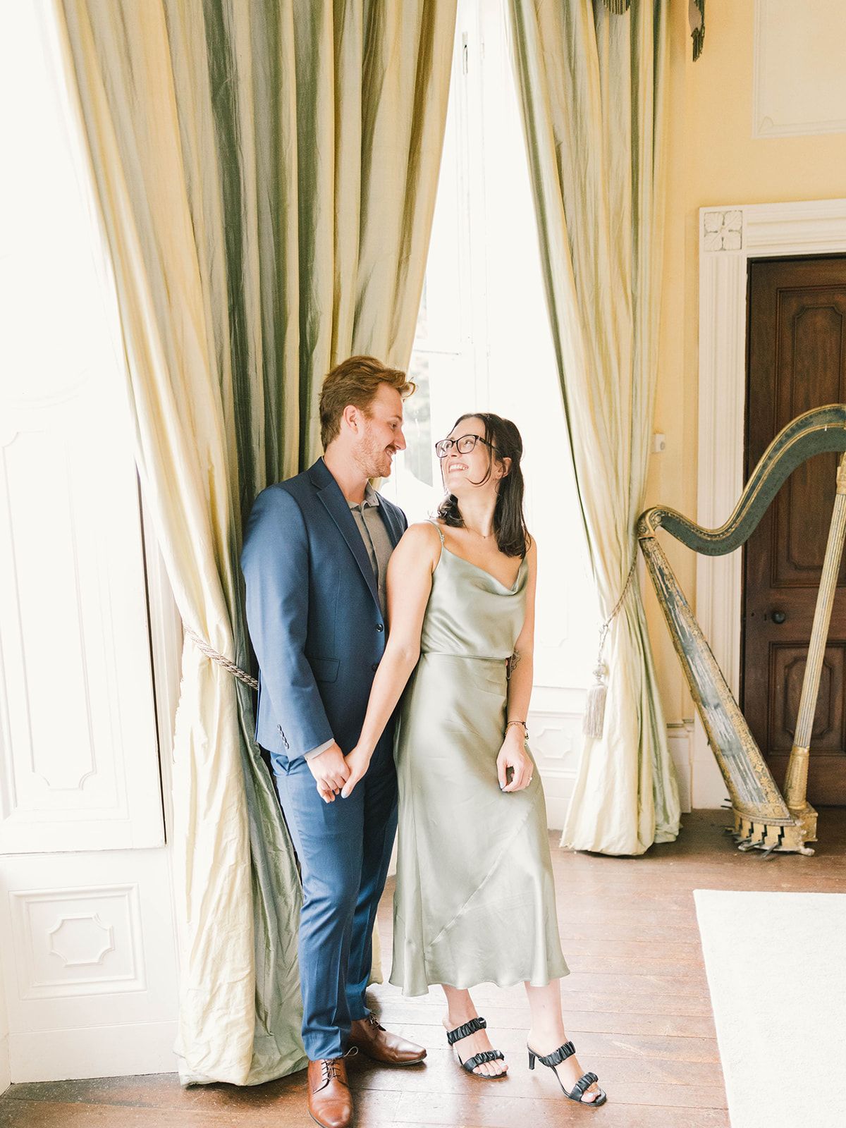 An elegant couple shares a romantic moment in a bright interior room with cream curtains and a decorative harp.