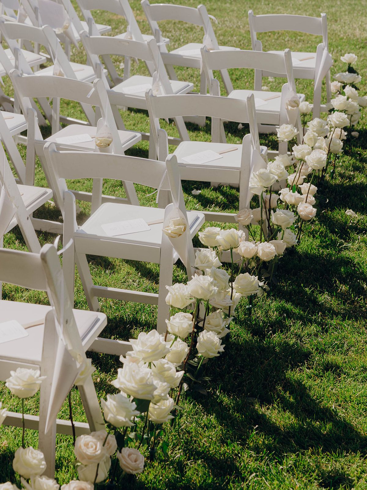 White wooden folding chairs on grass lined with elegant white roses for an outdoor wedding ceremony.