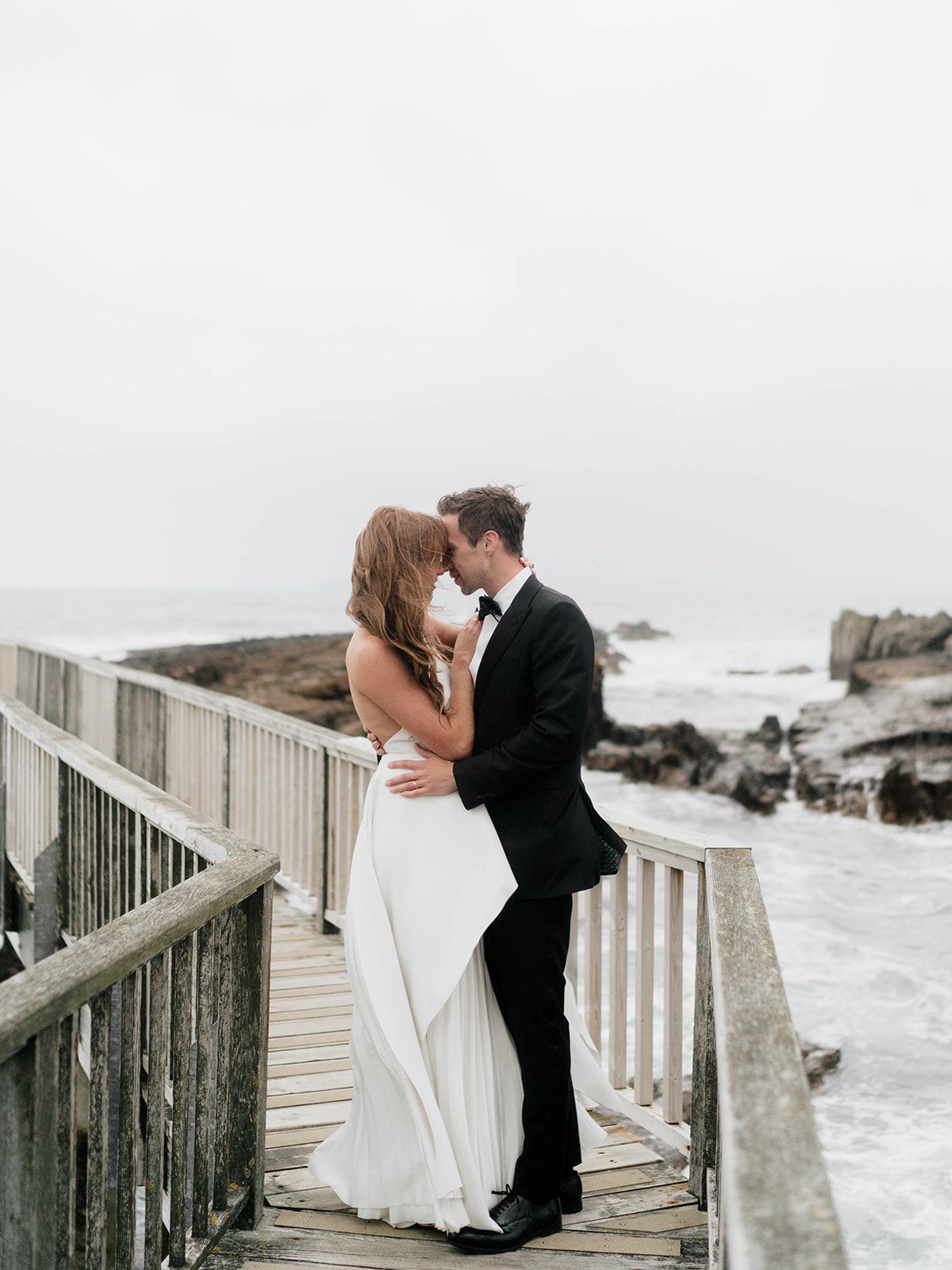 A bride and groom embrace on a wooden walkway overlooking a rocky coastline.