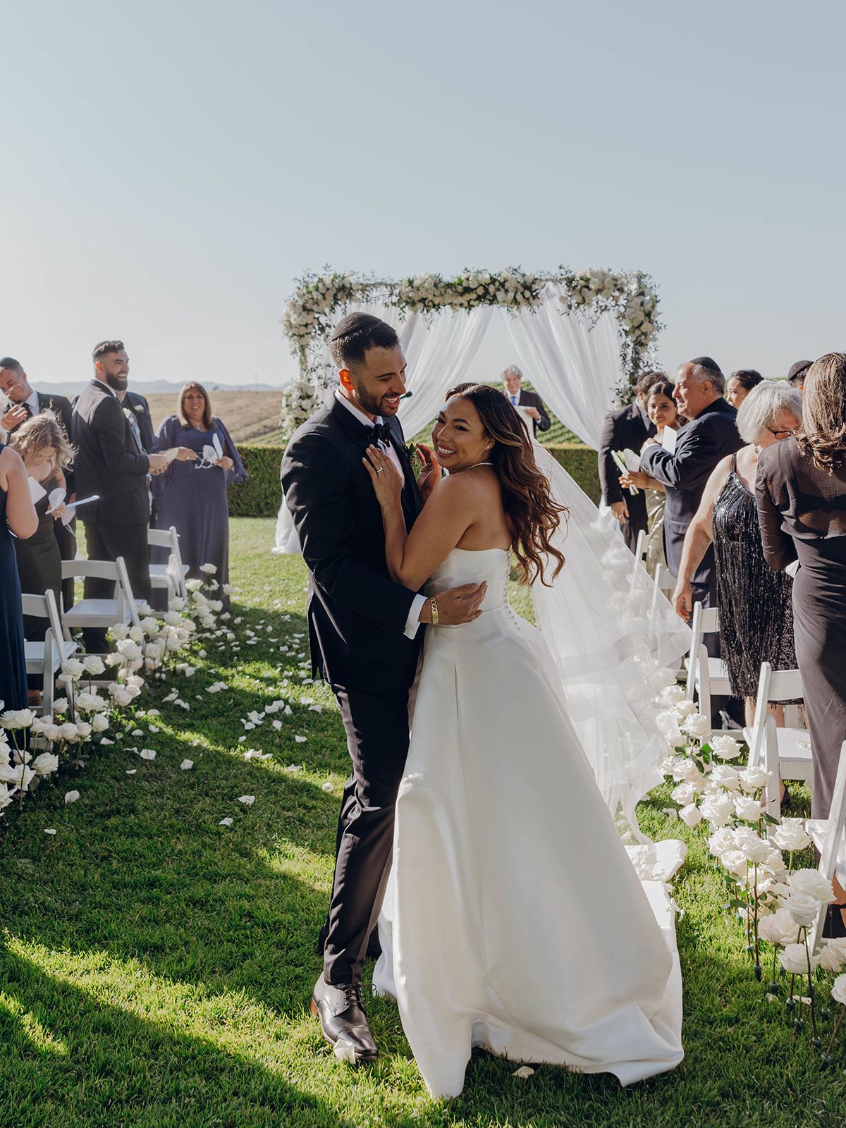 A joyful wedding couple embraces outdoors under a floral arch with guests looking on during a sunny ceremony.