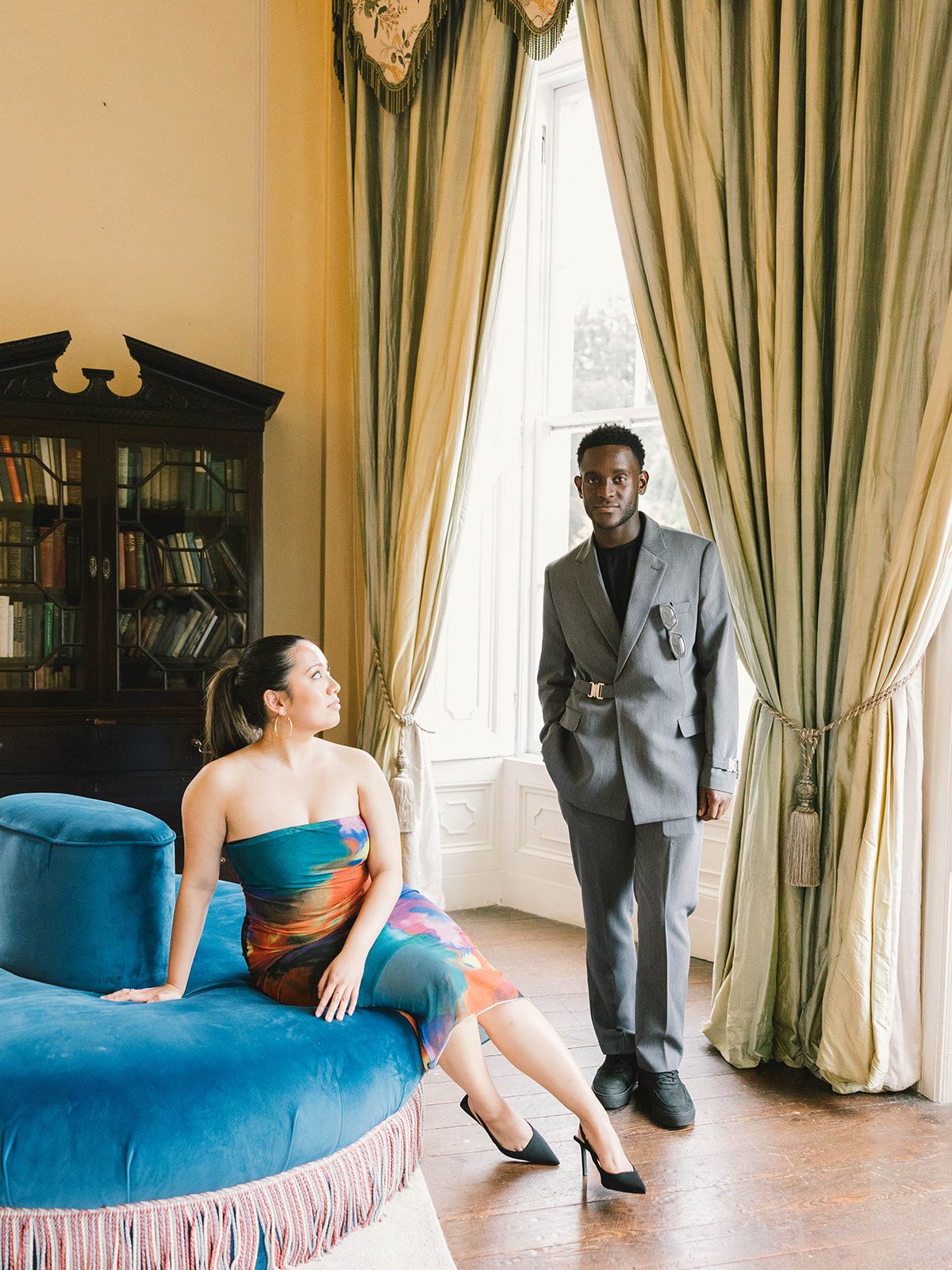 Two people in a grand room with tall windows and olive green curtains pose near a plush blue velvet chaise lounge.