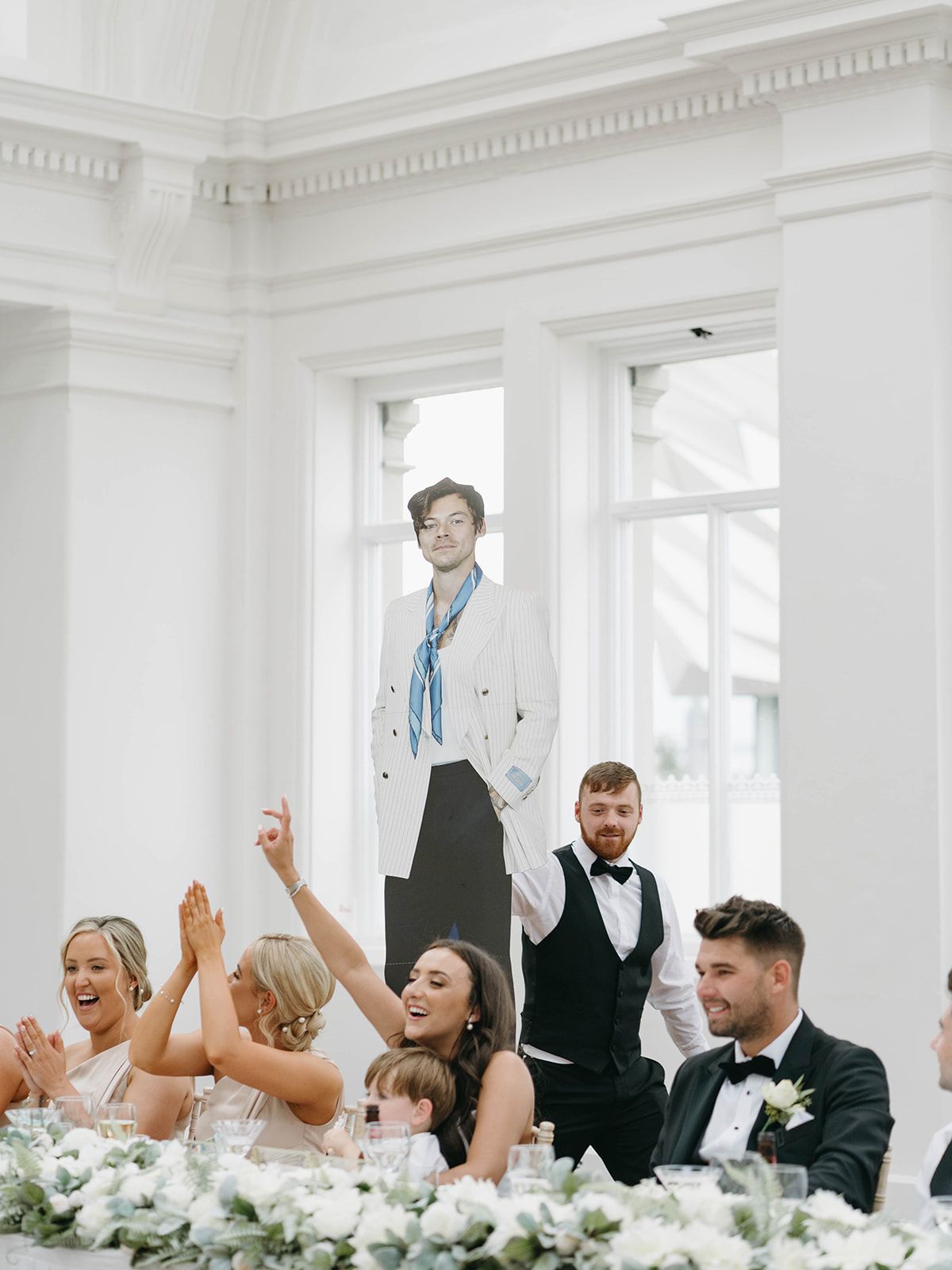 Wedding party celebrates joyfully at head table decorated with white floral arrangements in bright elegant venue.