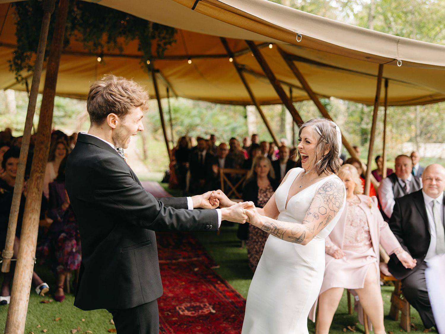 Couple shares their first dance under a tent canopy on a red carpet surrounded by wedding guests.