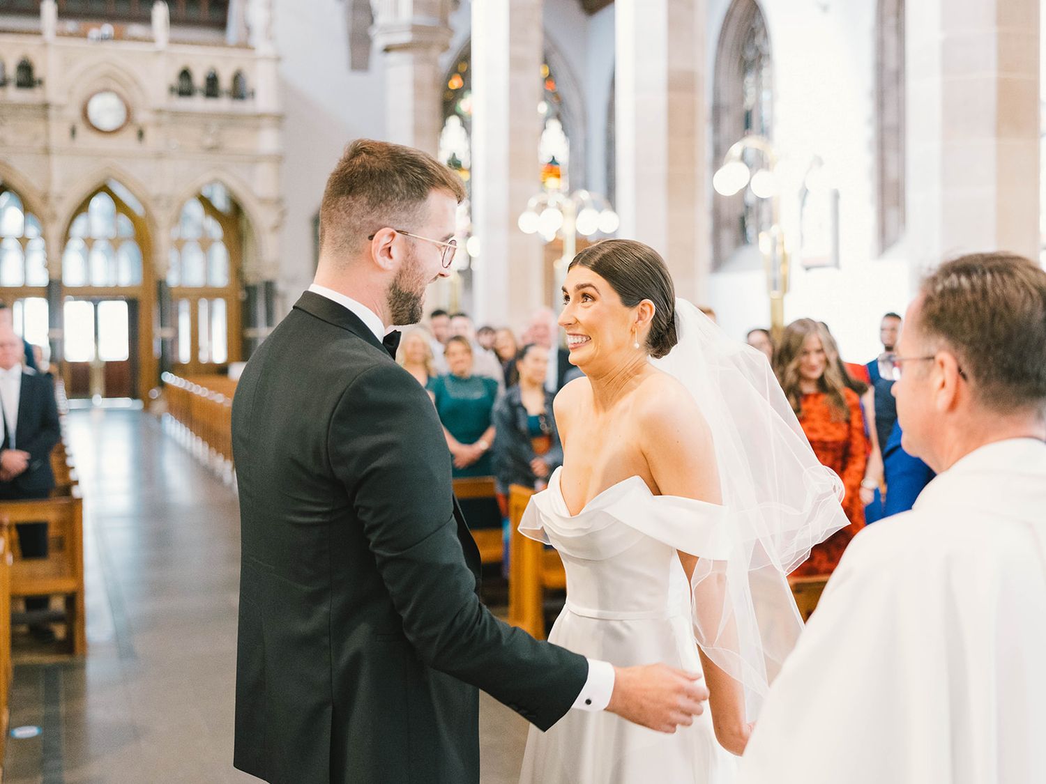 A couple exchanges vows during their wedding ceremony inside an ornate cathedral with arched windows and wooden pews.