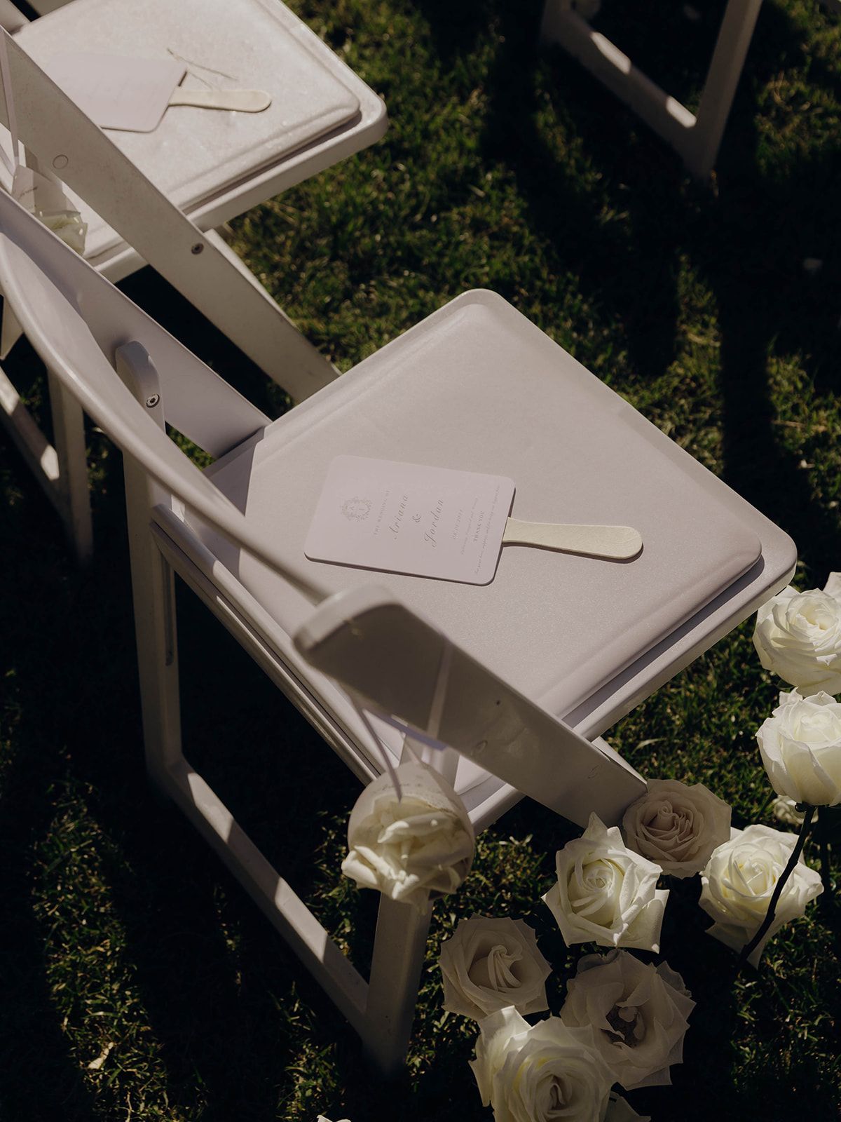White folding chairs decorated with white roses arranged for an outdoor wedding ceremony.