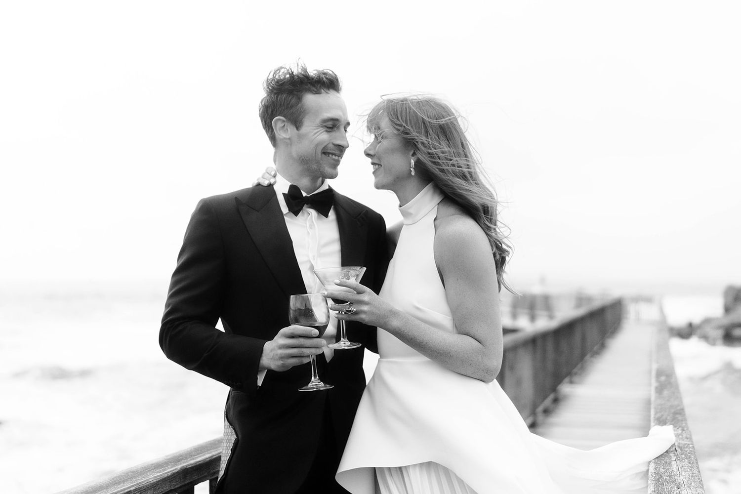 A couple in wedding attire shares champagne on a coastal boardwalk.