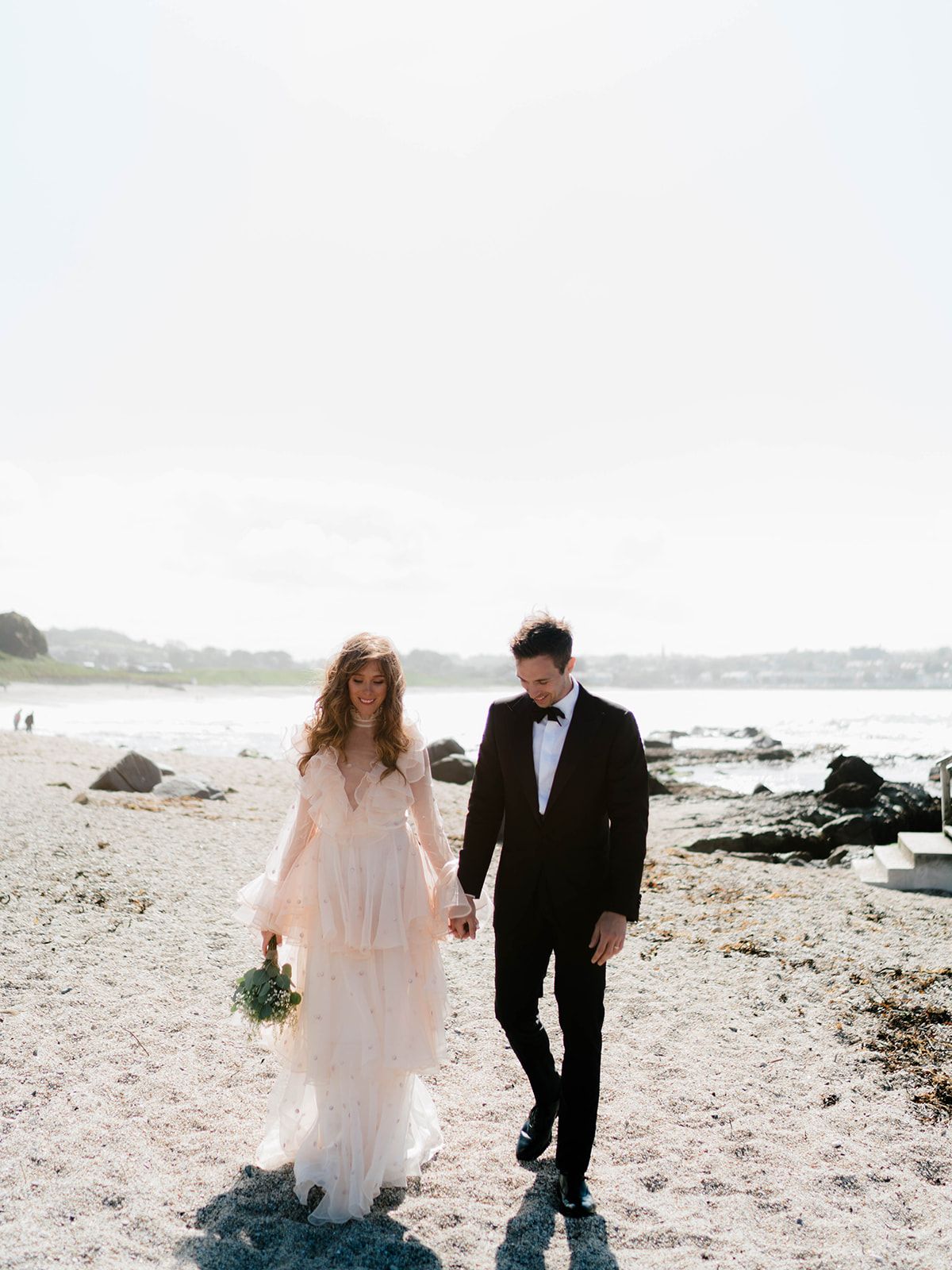 Wedding couple walking hand in hand along a rocky beach shoreline with misty scenery.