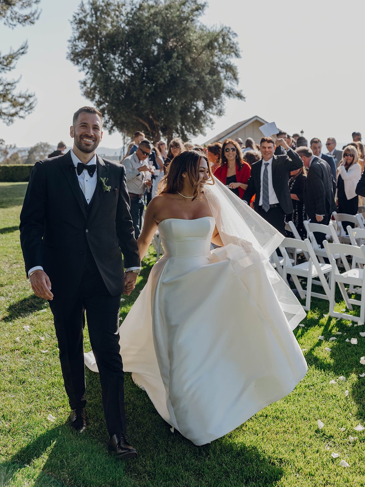 Bride and groom celebrate with guests at a sunny outdoor wedding ceremony with white chairs and a scenic backdrop.