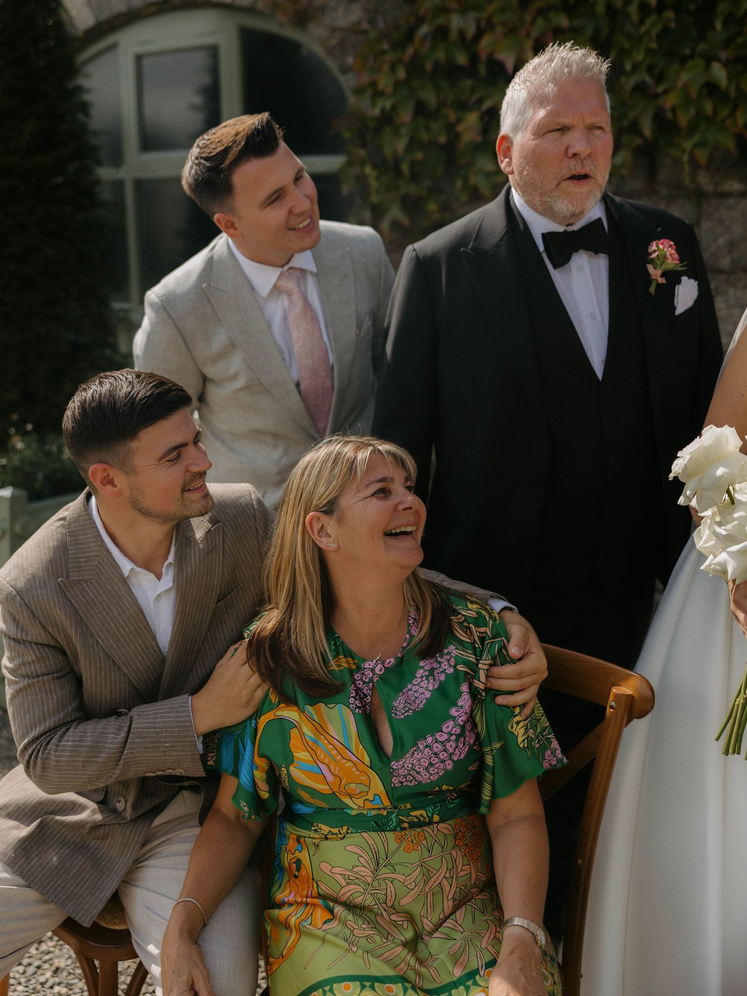 Wedding party members laugh joyfully together during outdoor celebration on a sunny day.