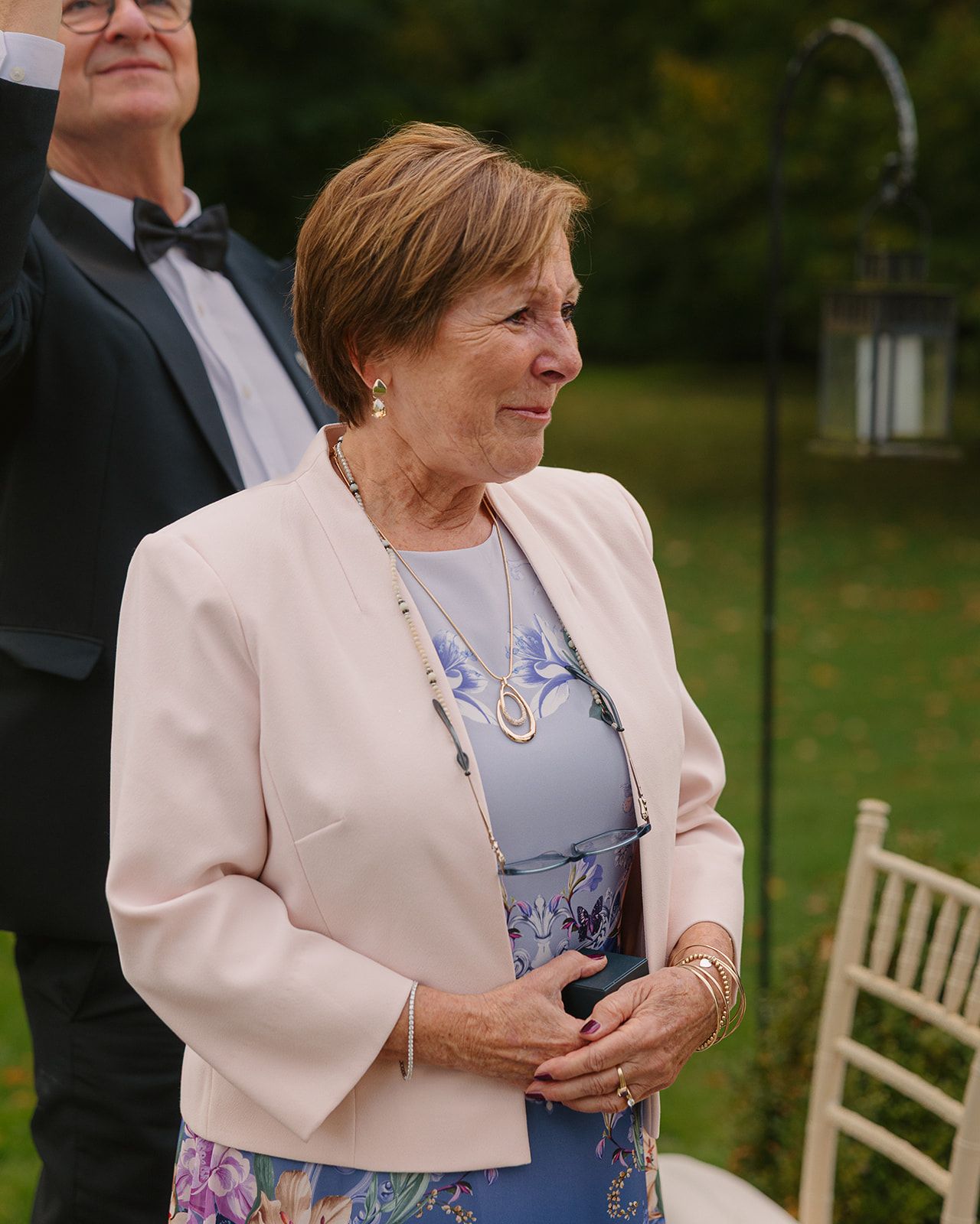 A person wearing a light pink blazer and floral dress stands outdoors near wooden chairs during an event.